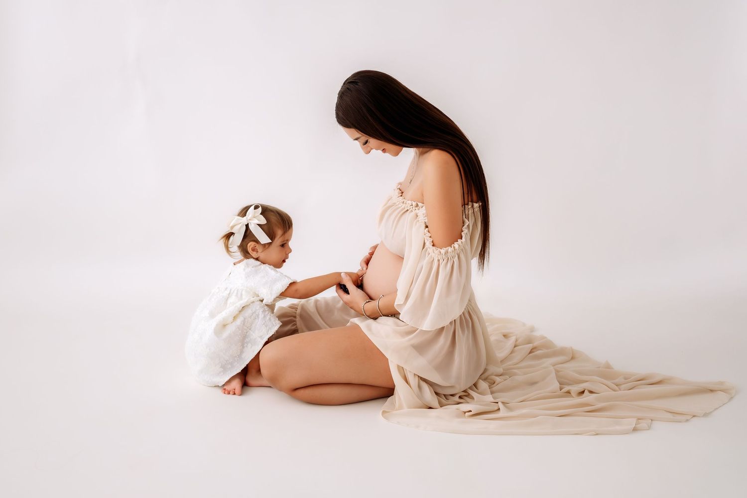 Toddler girl touching pregnant mothers belly during maternity photoshoot on white background.