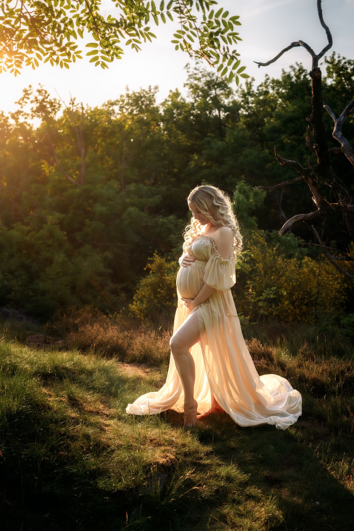 Pregnant woman in sheer peach off-shoulder gown holds belly in sunlit forest clearing at golden hour.