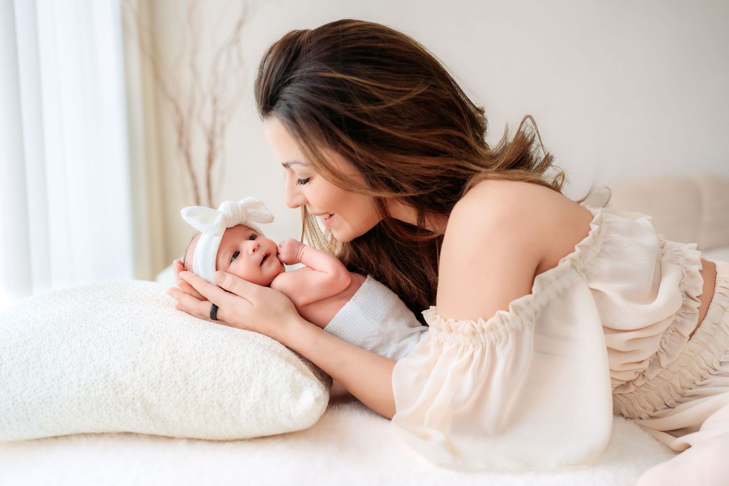 Young mother in off-shoulder dress lovingly gazing at her newborn baby on bed.