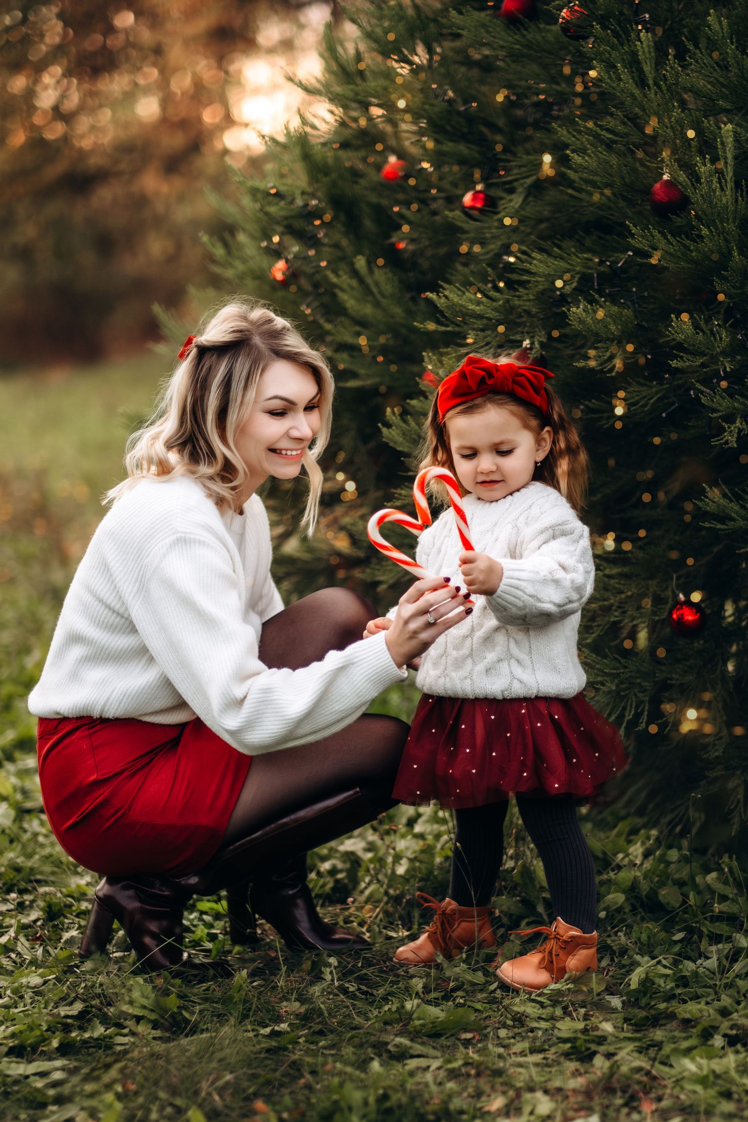 Mother and toddler daughter in matching red and white outfits holding candy canes by decorated Christmas tree outdoors.