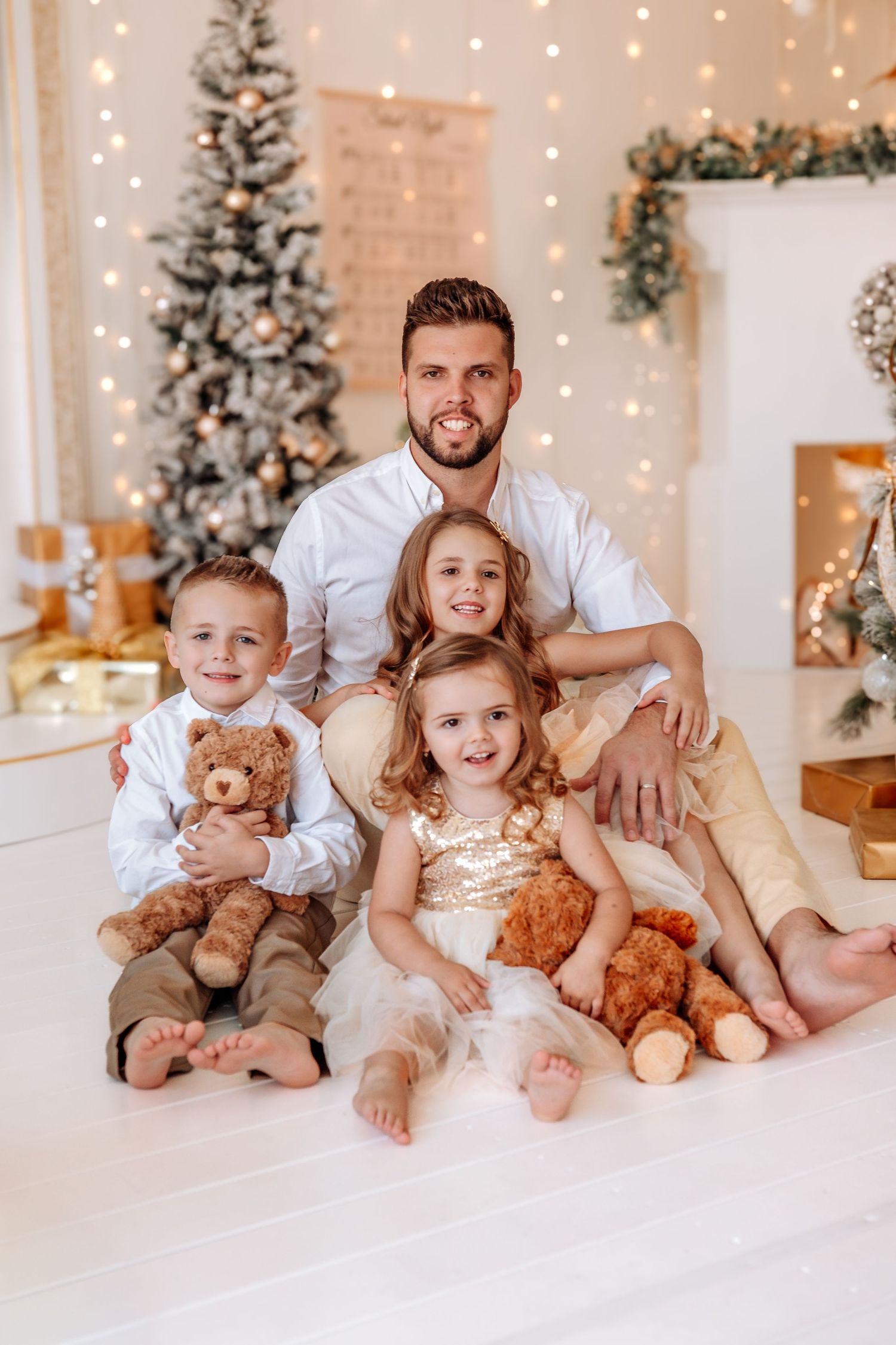 Father poses with three young children holding teddy bears beside a decorated Christmas tree and fireplace.