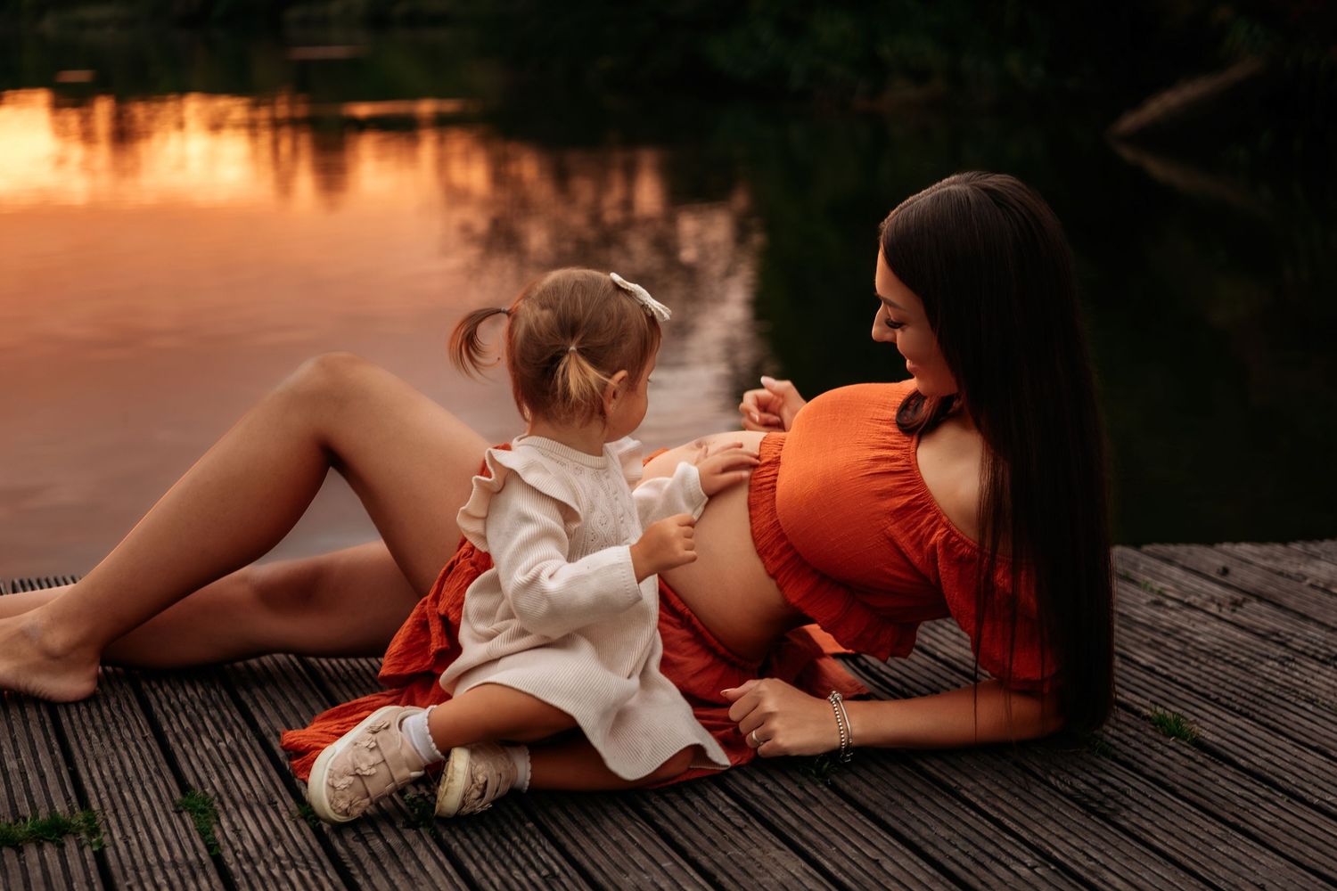 Pregnant woman in red crop top reclines on dock while toddler daughter touches her baby bump at sunset.