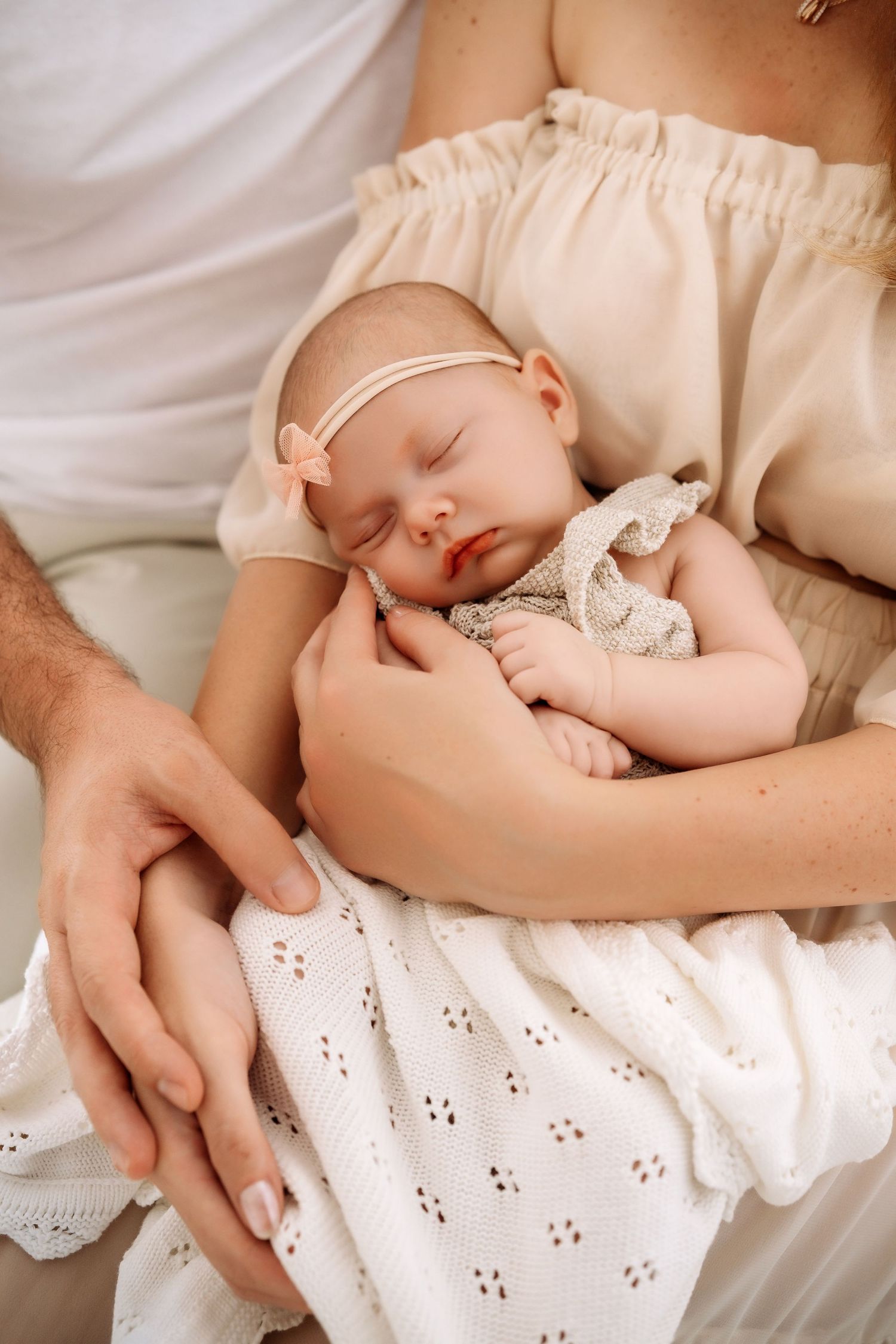 Parents hands cradling sleeping newborn baby in white blanket and pink headband together.