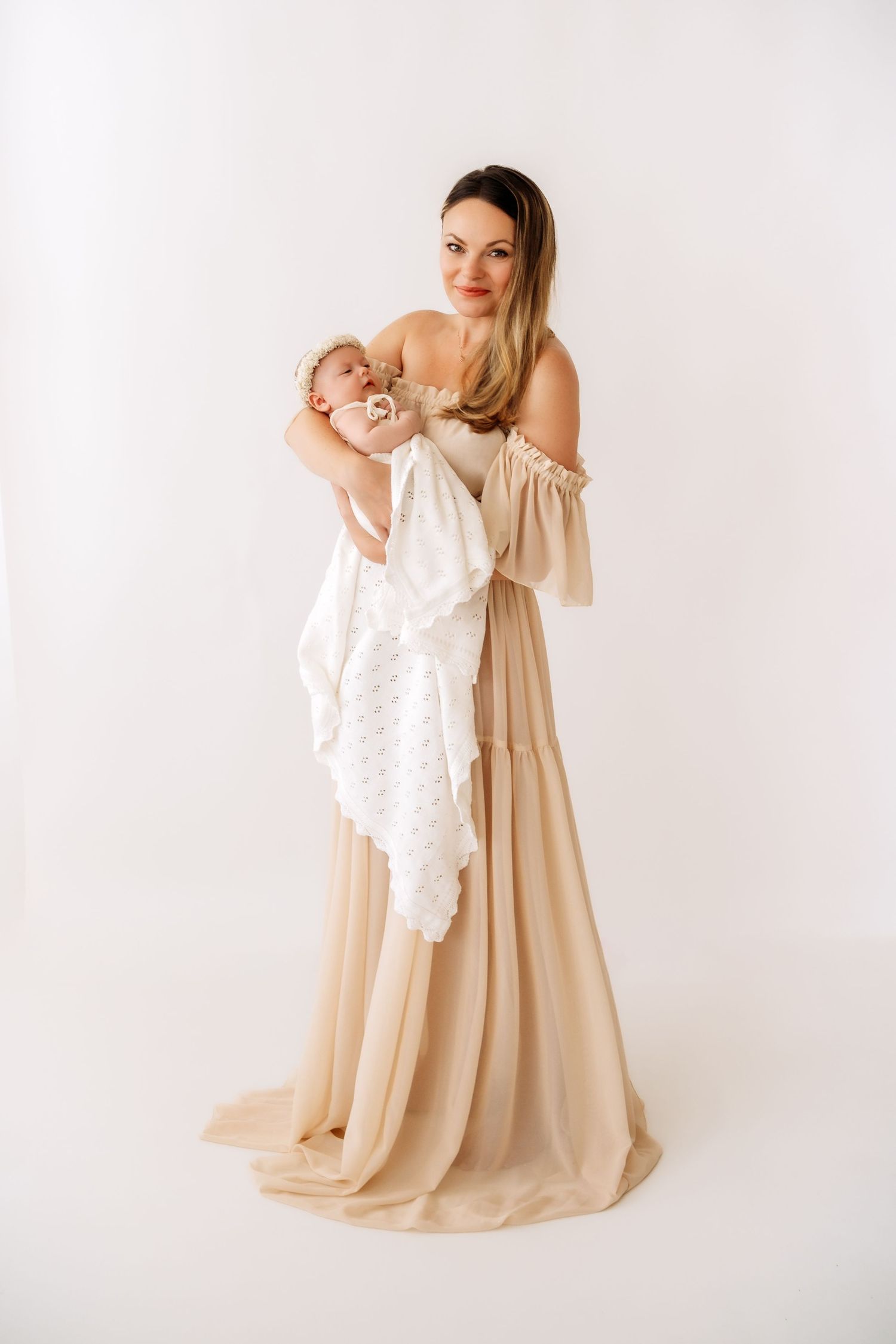 Elegant mother in flowing beige gown holds newborn wrapped in white lace blanket in studio.