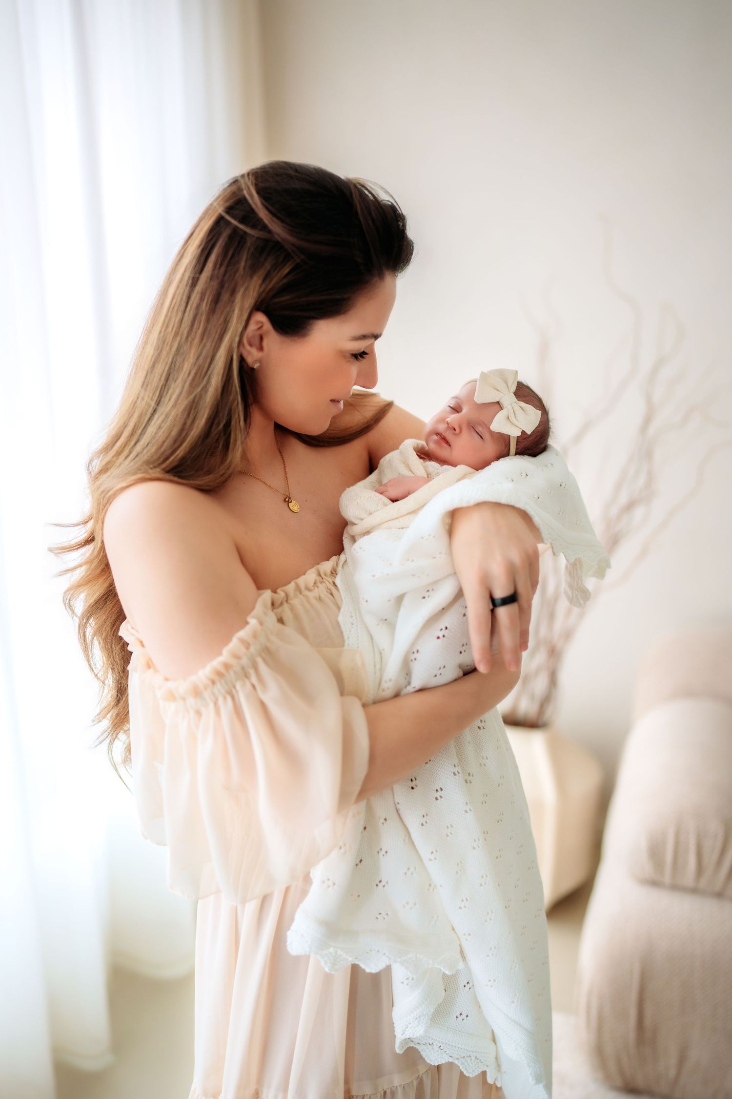 A woman in a blush off-shoulder dress lovingly gazes at her swaddled newborn baby girl indoors.