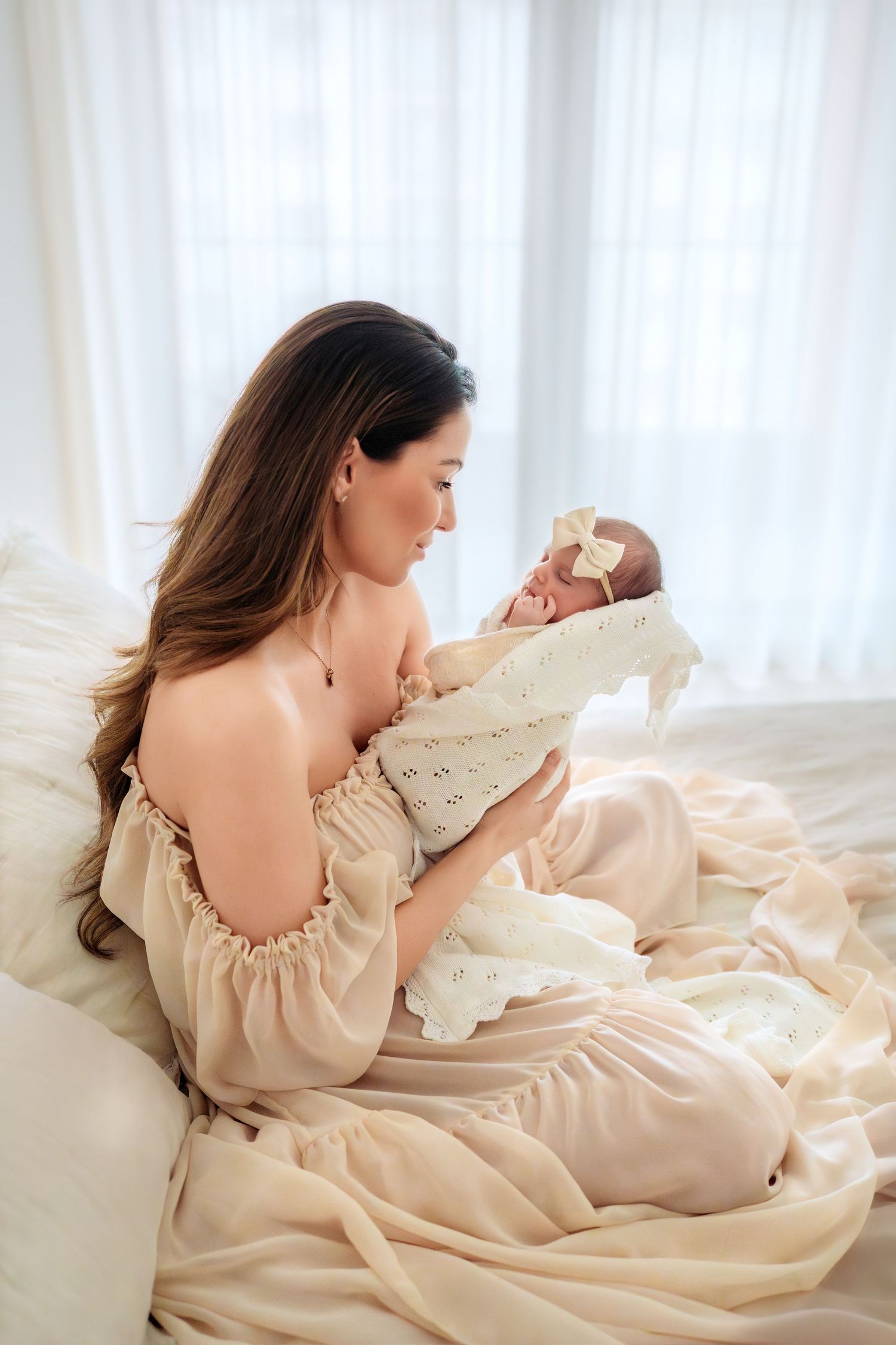 A dark-haired mother in a cream dress sits on a bed lovingly holding her swaddled newborn baby girl.