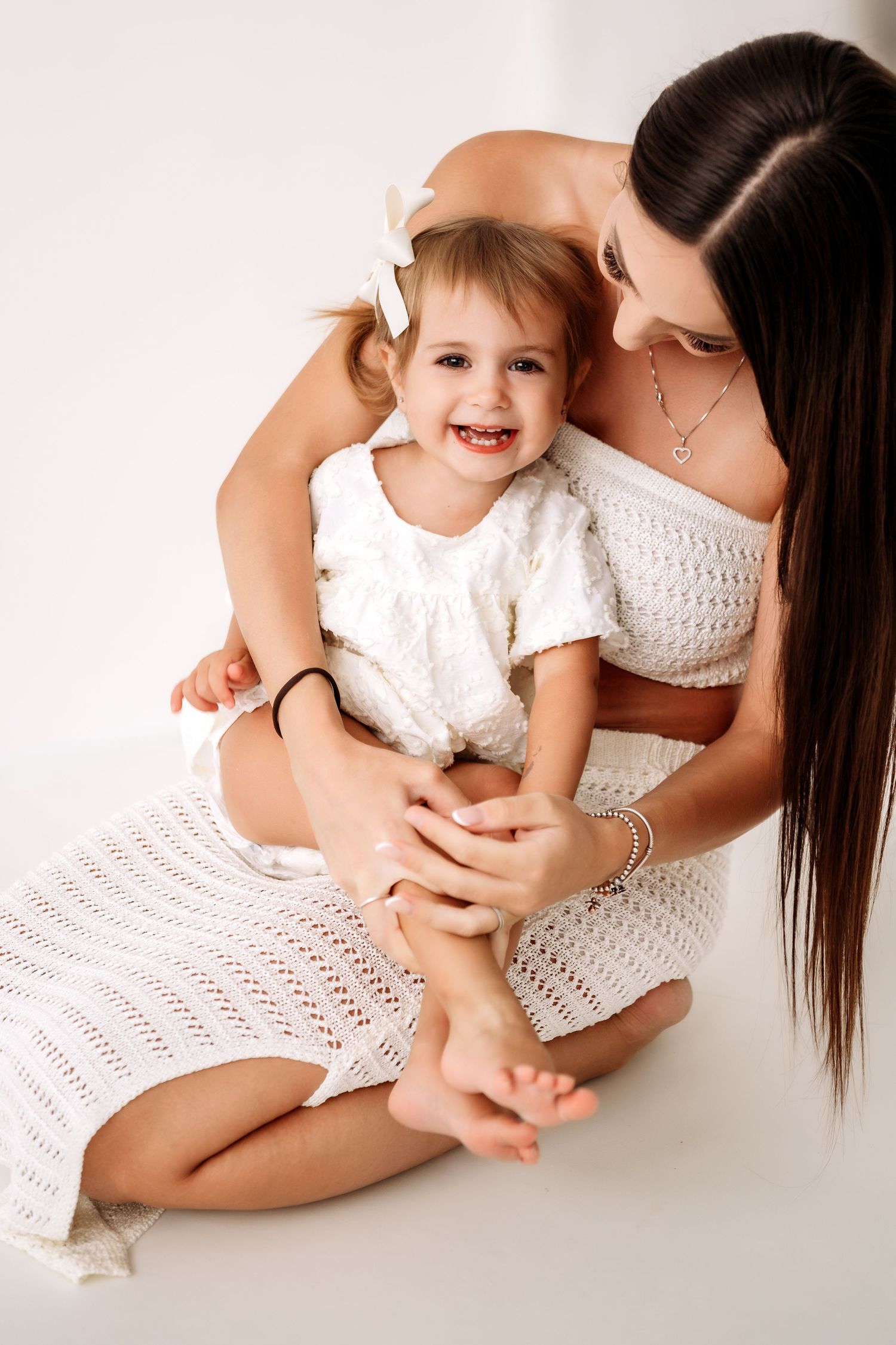 Pregnant mother in white knit dress sits on floor holding smiling toddler daughter in white dress.