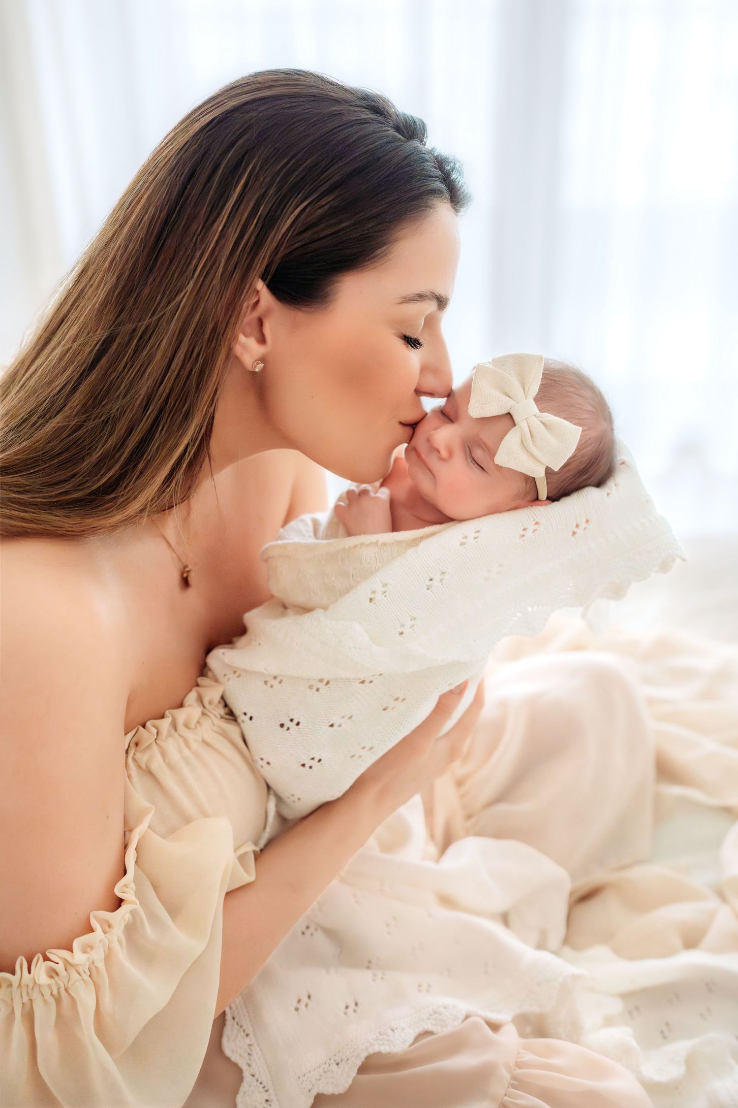 Loving mother in cream dress gently kissing newborn baby girl wearing white bow headband.