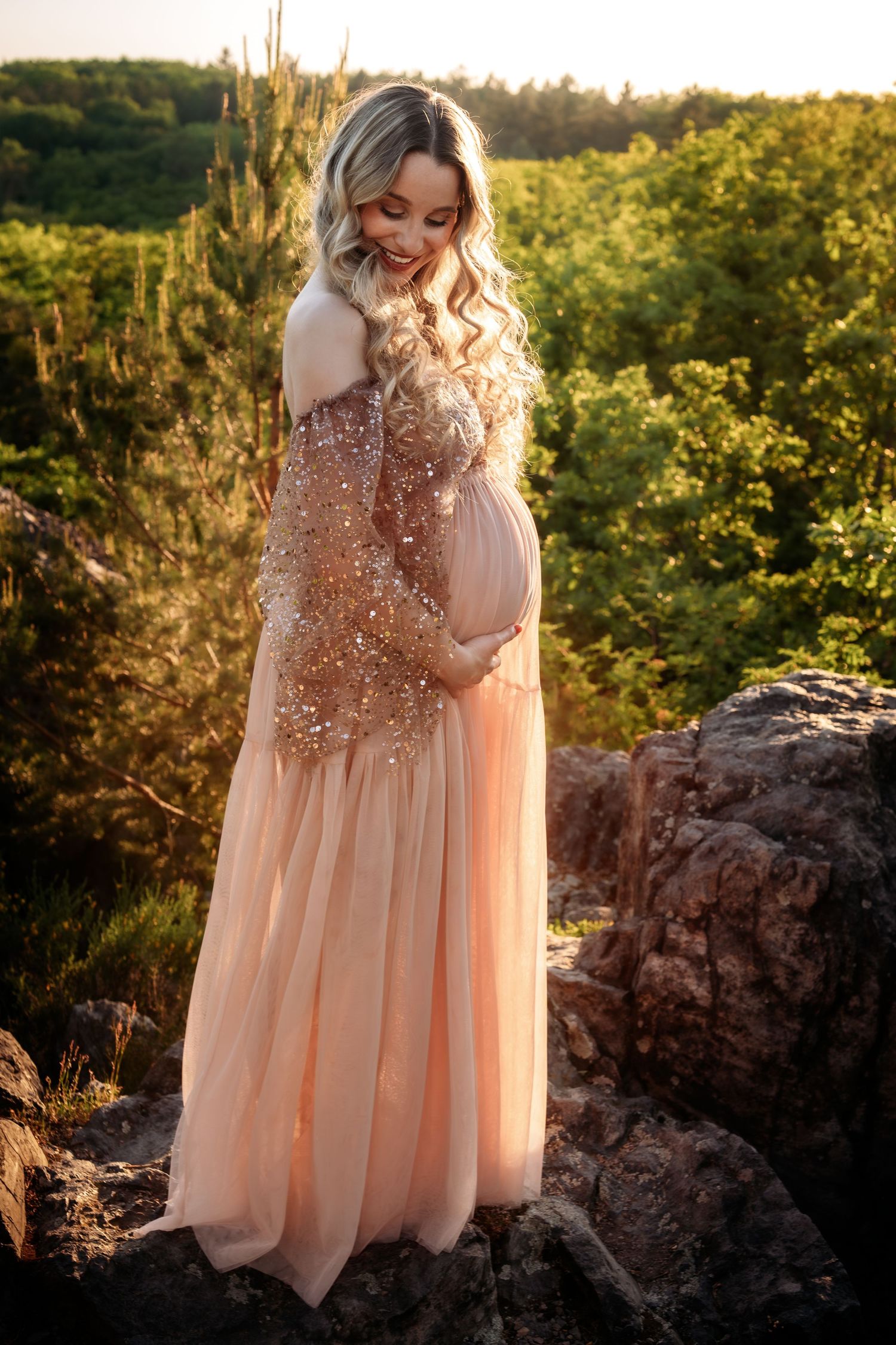 Pregnant woman in sequined peach gown cradles belly on rocky outcrop with green forest at golden sunset.