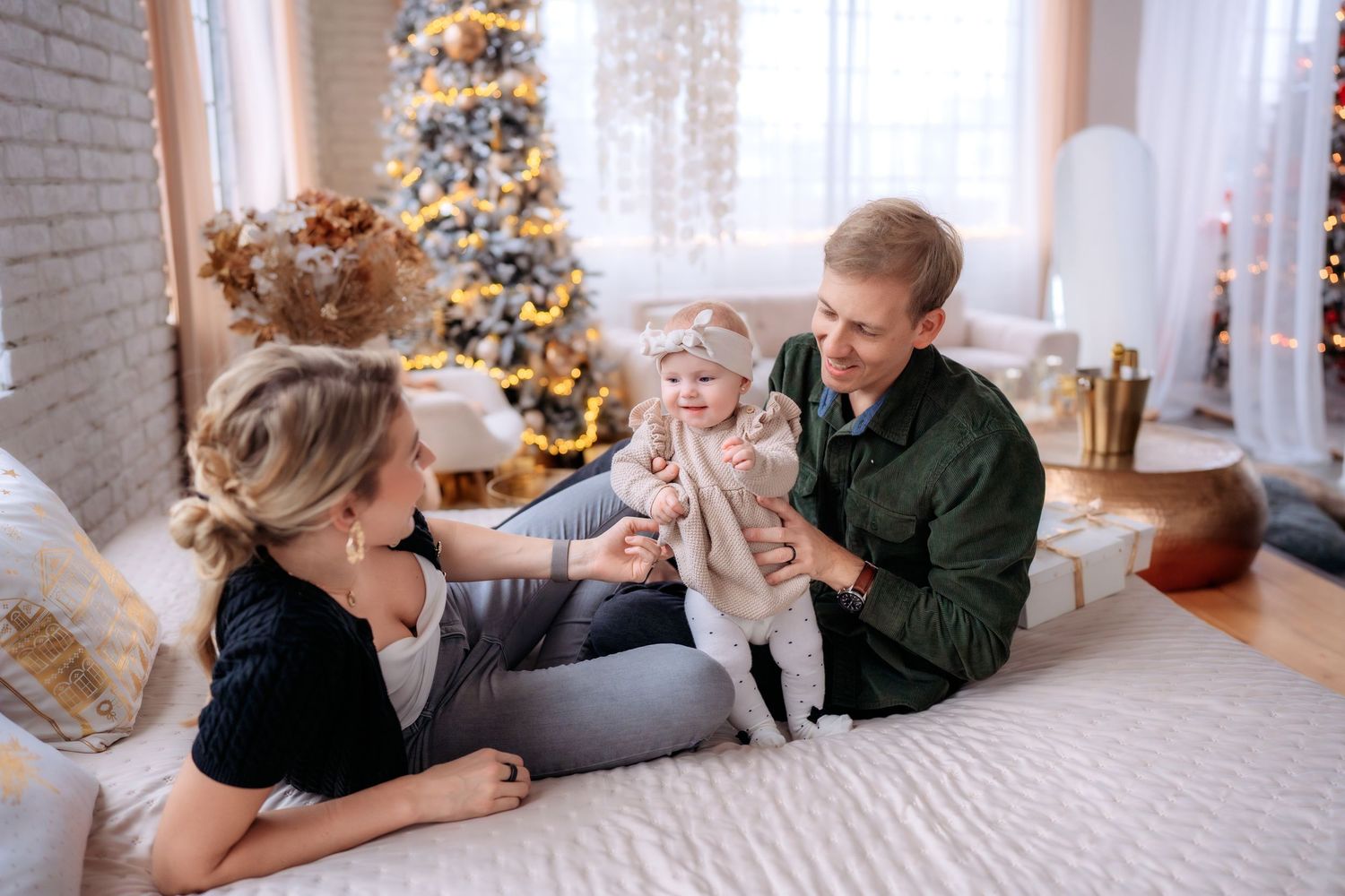 Young family with baby celebrating Christmas, sitting on bed near decorated tree in cozy living room.