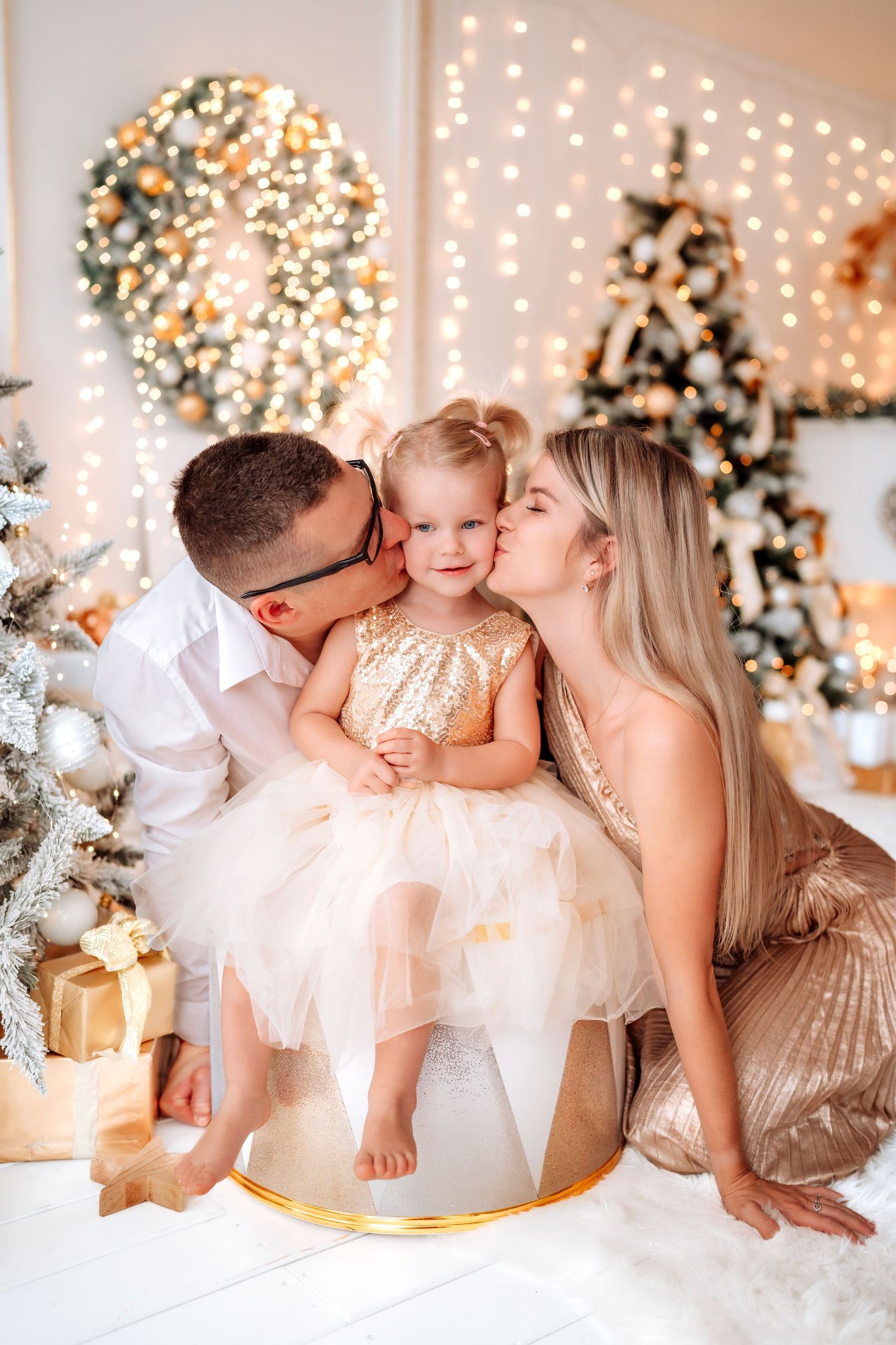 Family Christmas photo with parents kissing toddler girl in gold dress beside decorated trees and warm glowing lights.