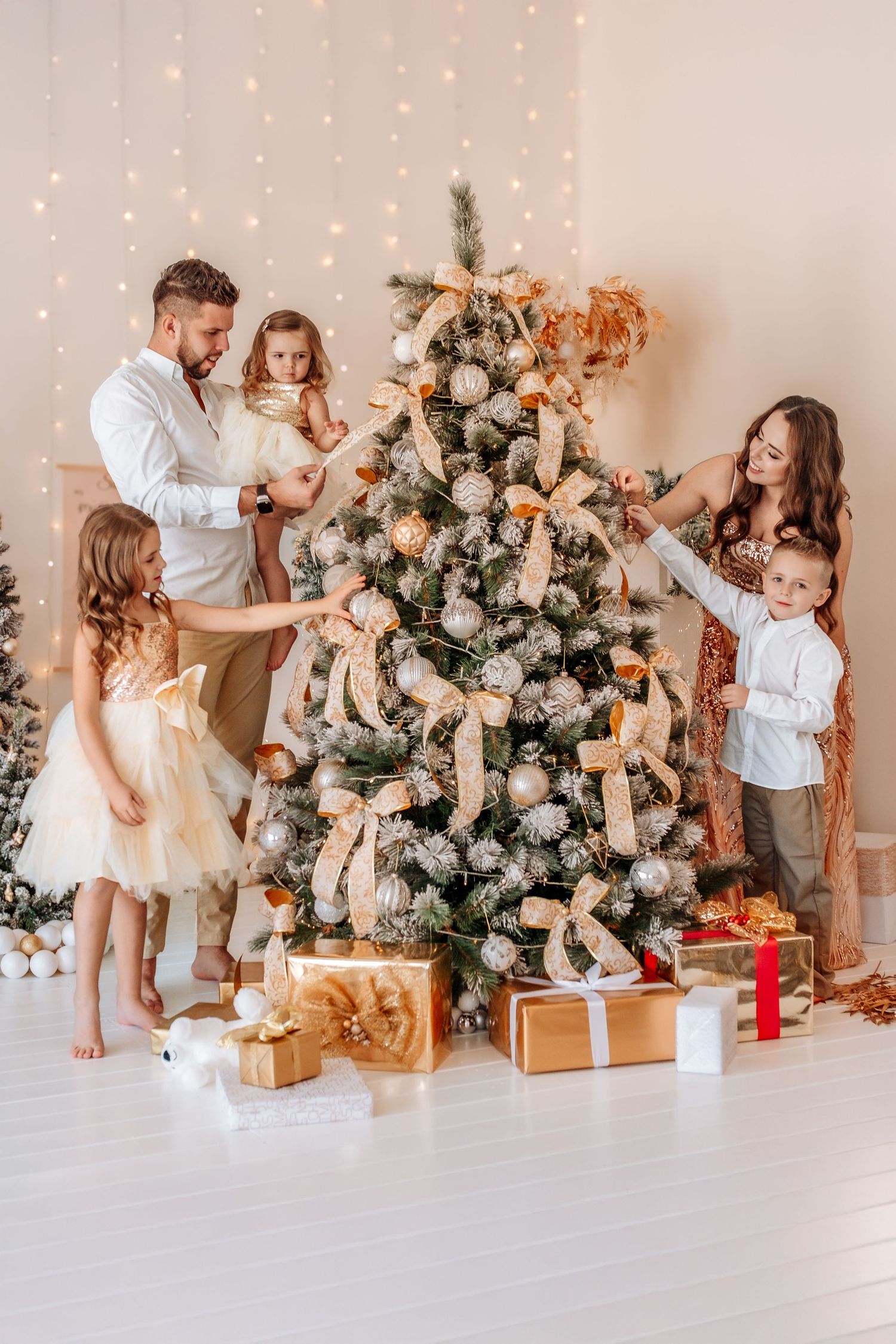 Family decorating a Christmas tree with gold ornaments and bows, surrounded by wrapped gifts in a bright festive room.