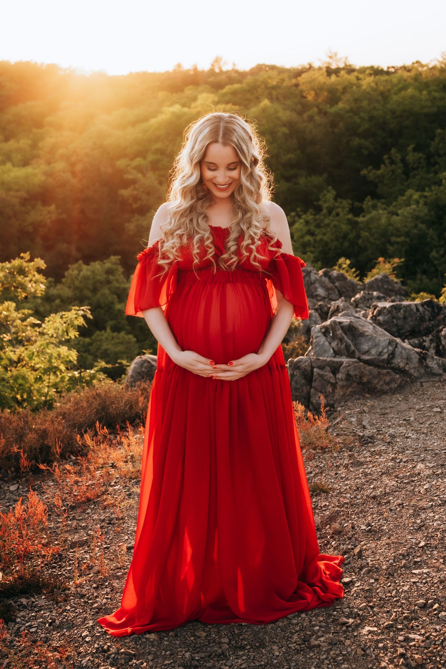 Pregnant woman in red off-shoulder gown standing outdoors at golden sunset with rocky landscape and green trees.