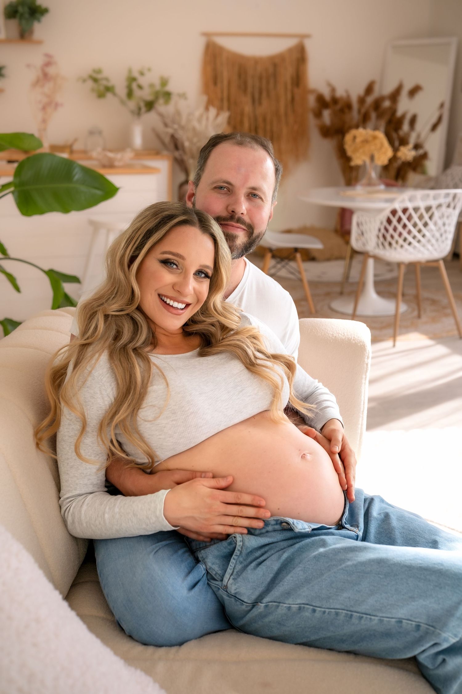 Joyful pregnant woman with long blonde hair sitting on couch, partner embracing her from behind, hands on baby bump.