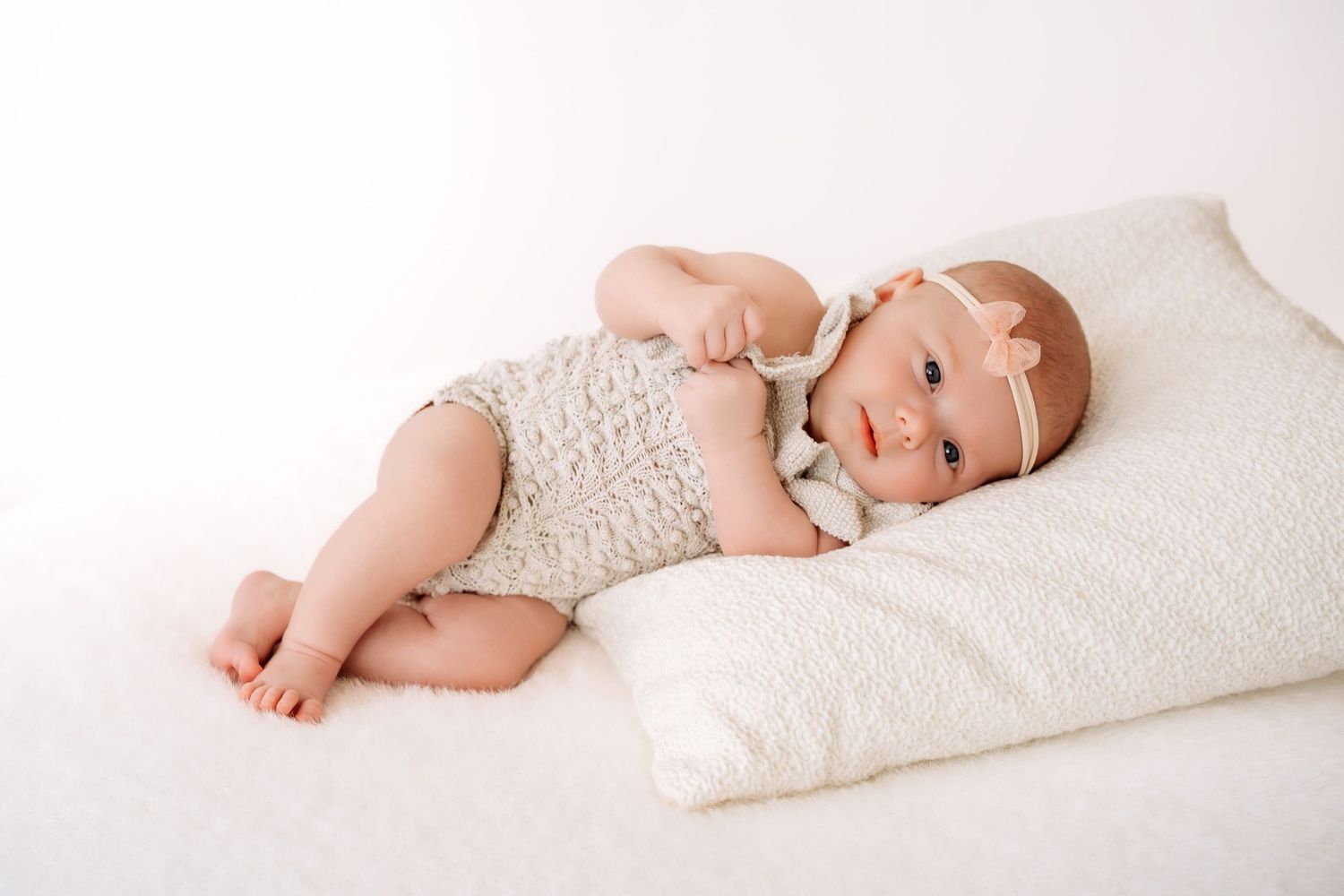 Sweet baby girl in cream knit romper resting on white pillow with bare feet.