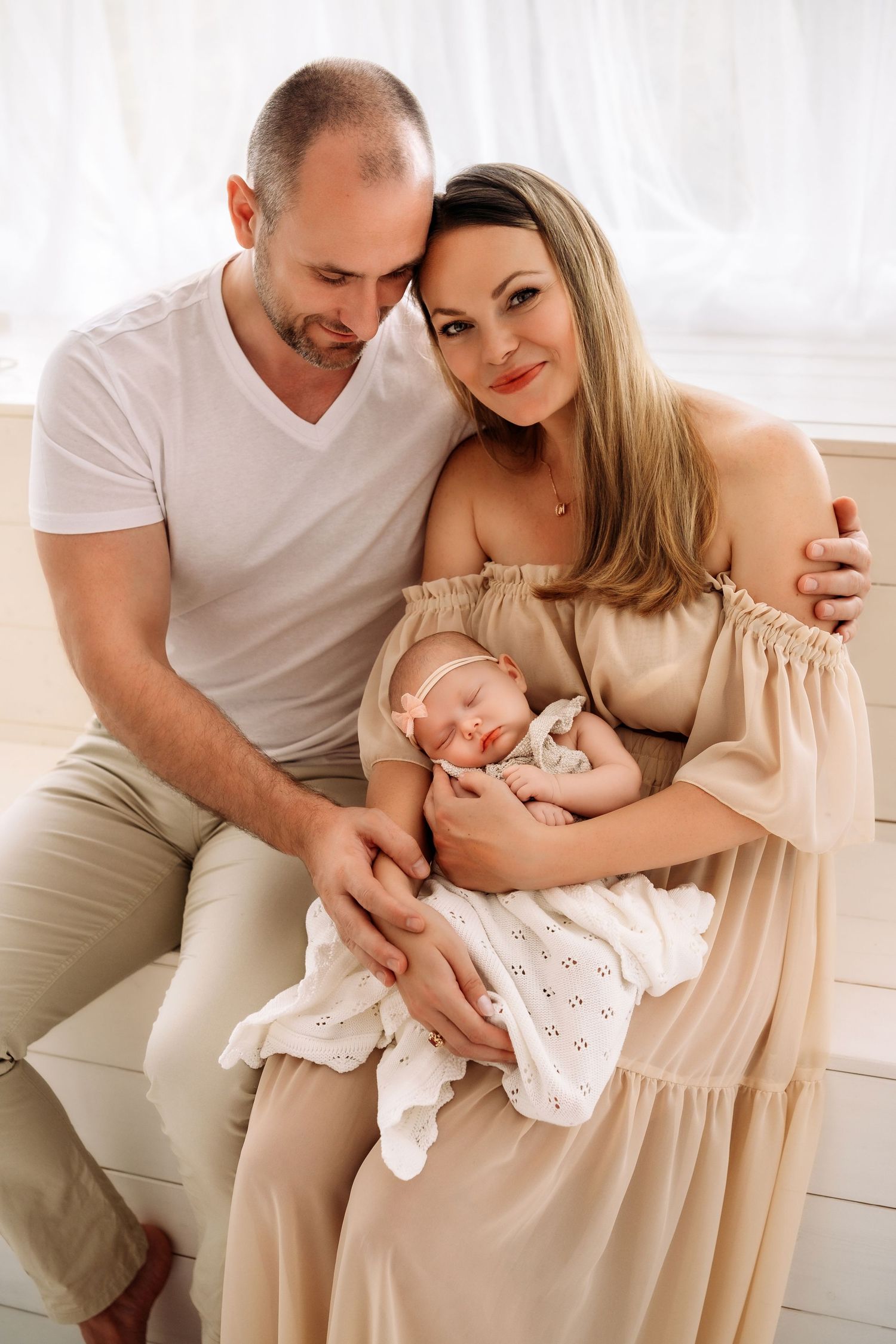 Happy parents sitting together holding their sleeping newborn baby girl in white blanket.