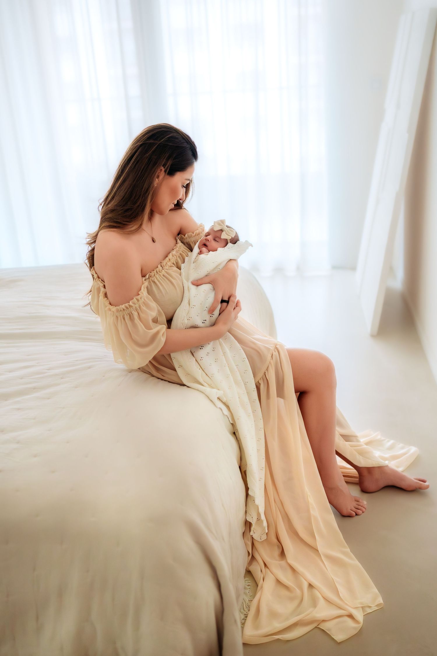 A mother in a beige off-shoulder gown sits on a bed cradling her newborn baby near a bright window.