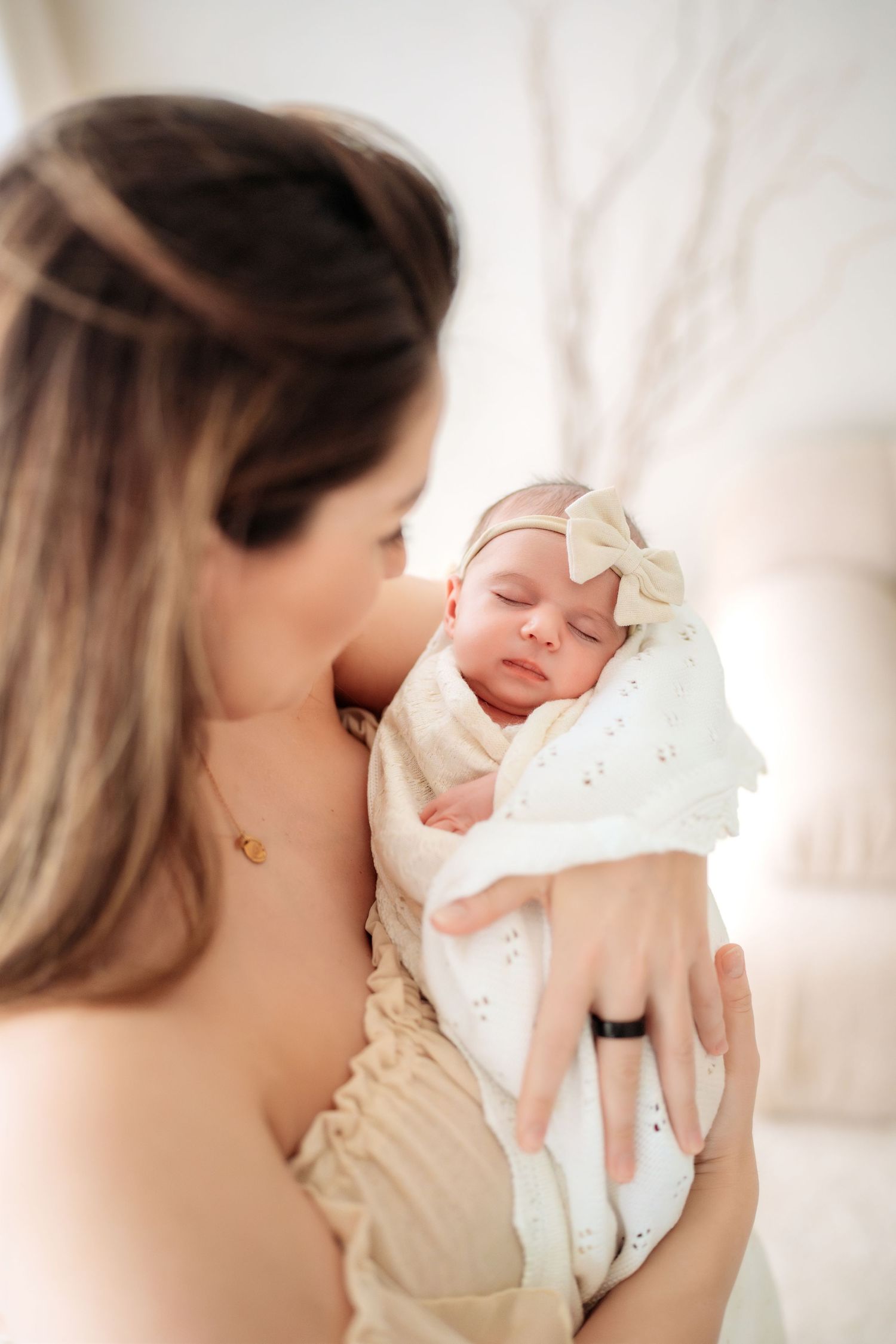 Mother holding swaddled newborn baby girl with bow headband close to her chest in bright room.