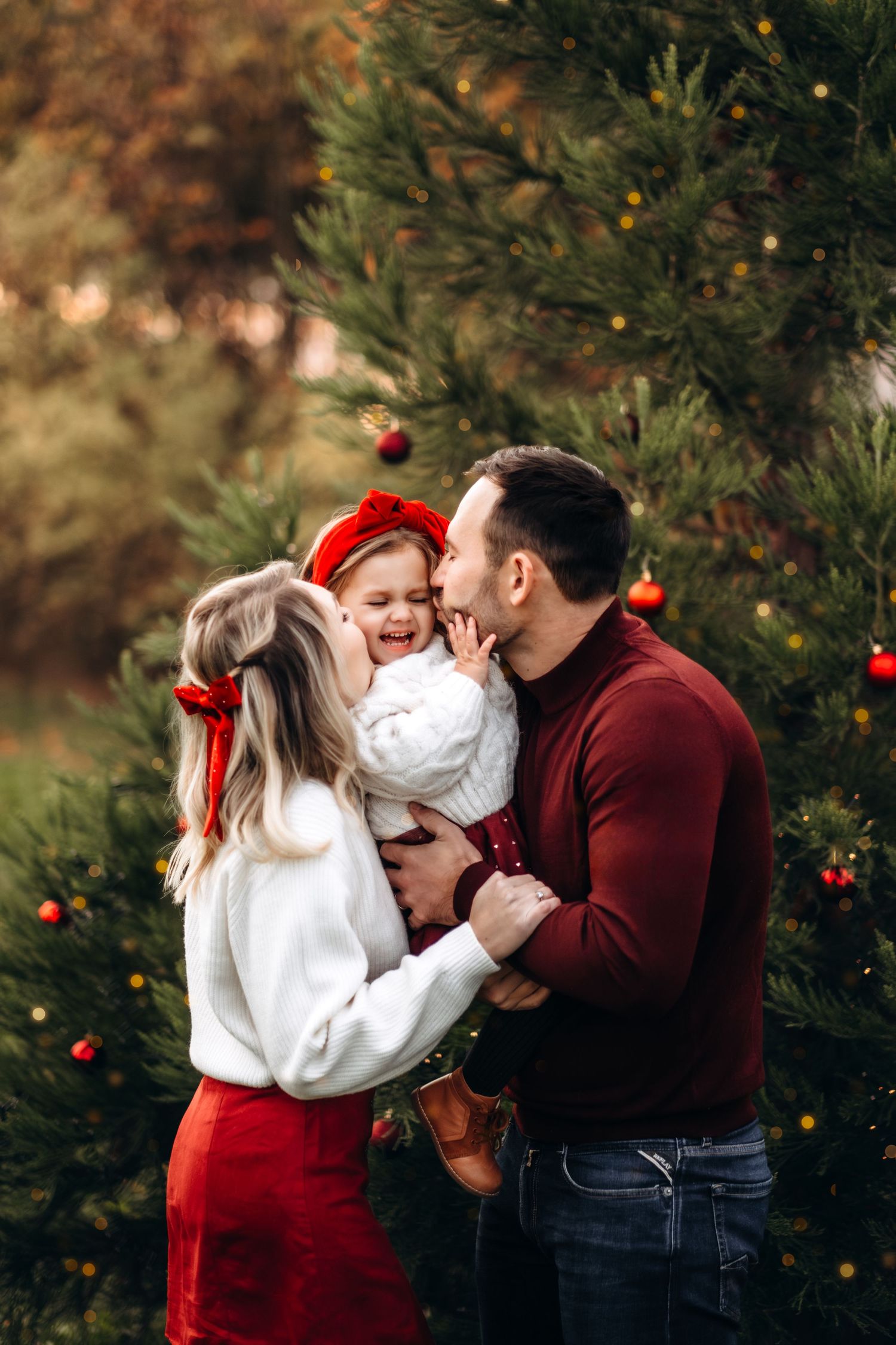 A happy family with parents kissing their laughing toddler girl outdoors near decorated Christmas trees.