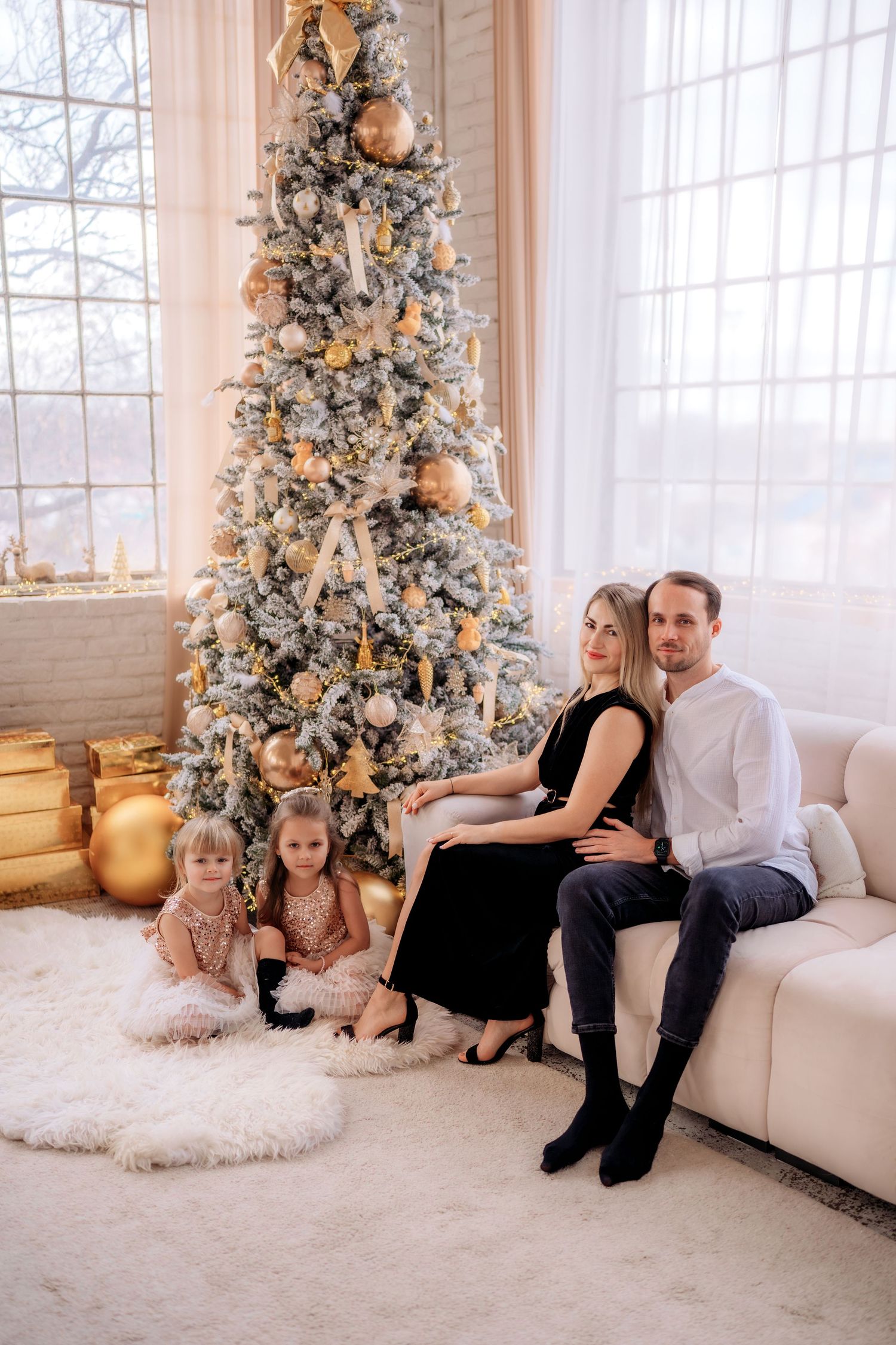 Family of four posing by a decorated Christmas tree with gold ornaments, two girls sitting on floor, parents on white sofa.