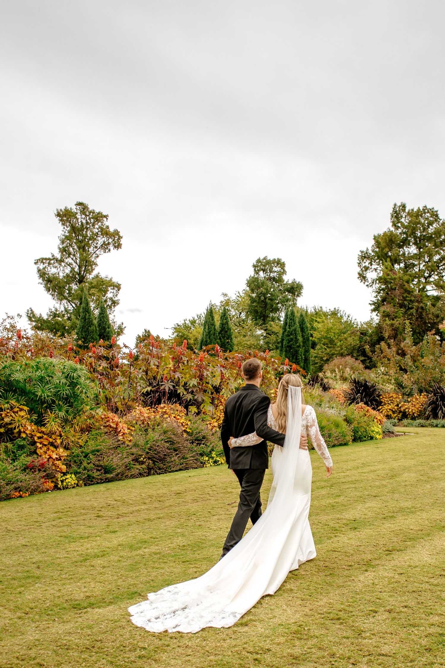 Bride and groom walk hand-in-hand away from camera through vibrant autumn garden landscape.