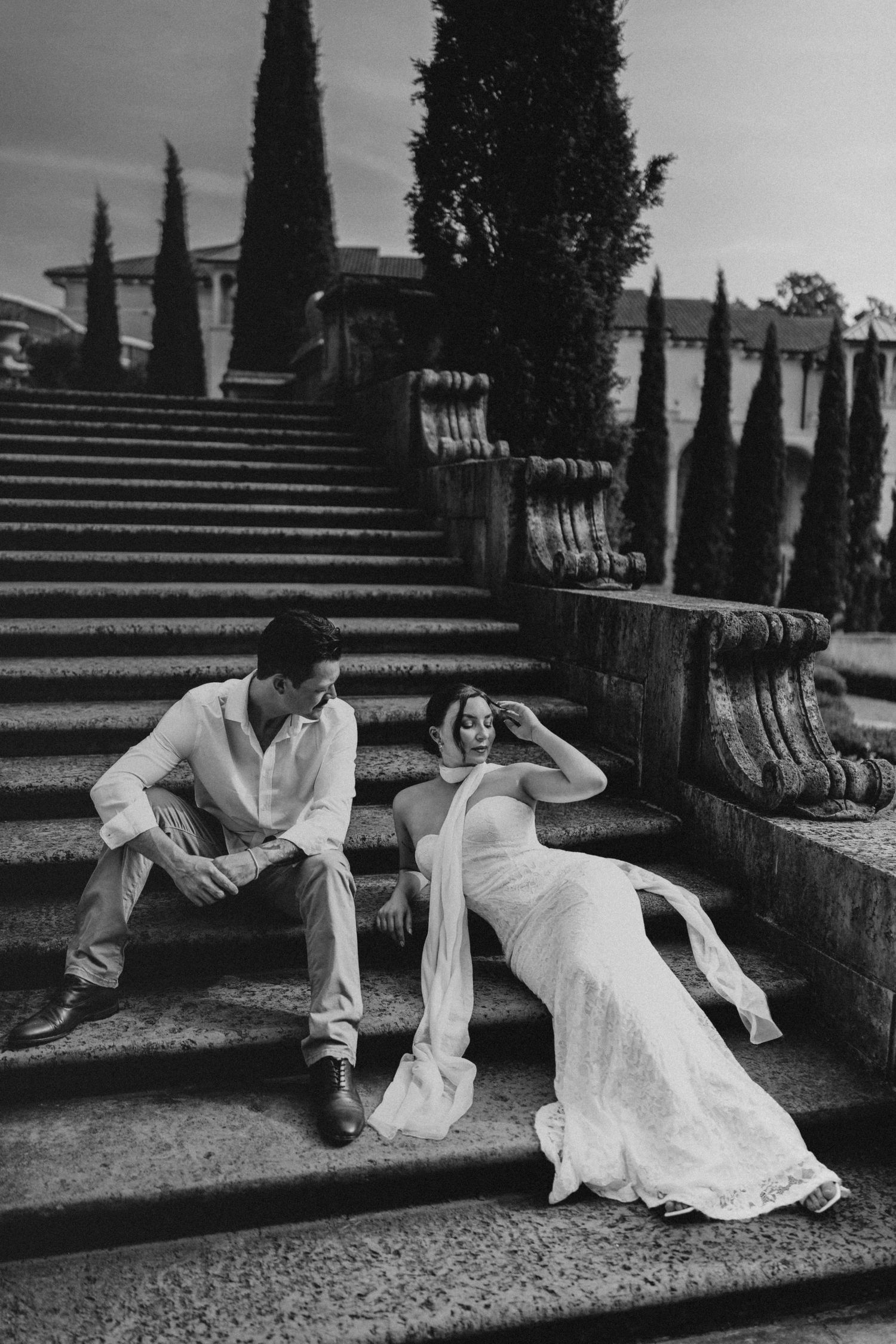 Bride in white gown and groom in white shirt sit on stone steps with cypress trees behind them.