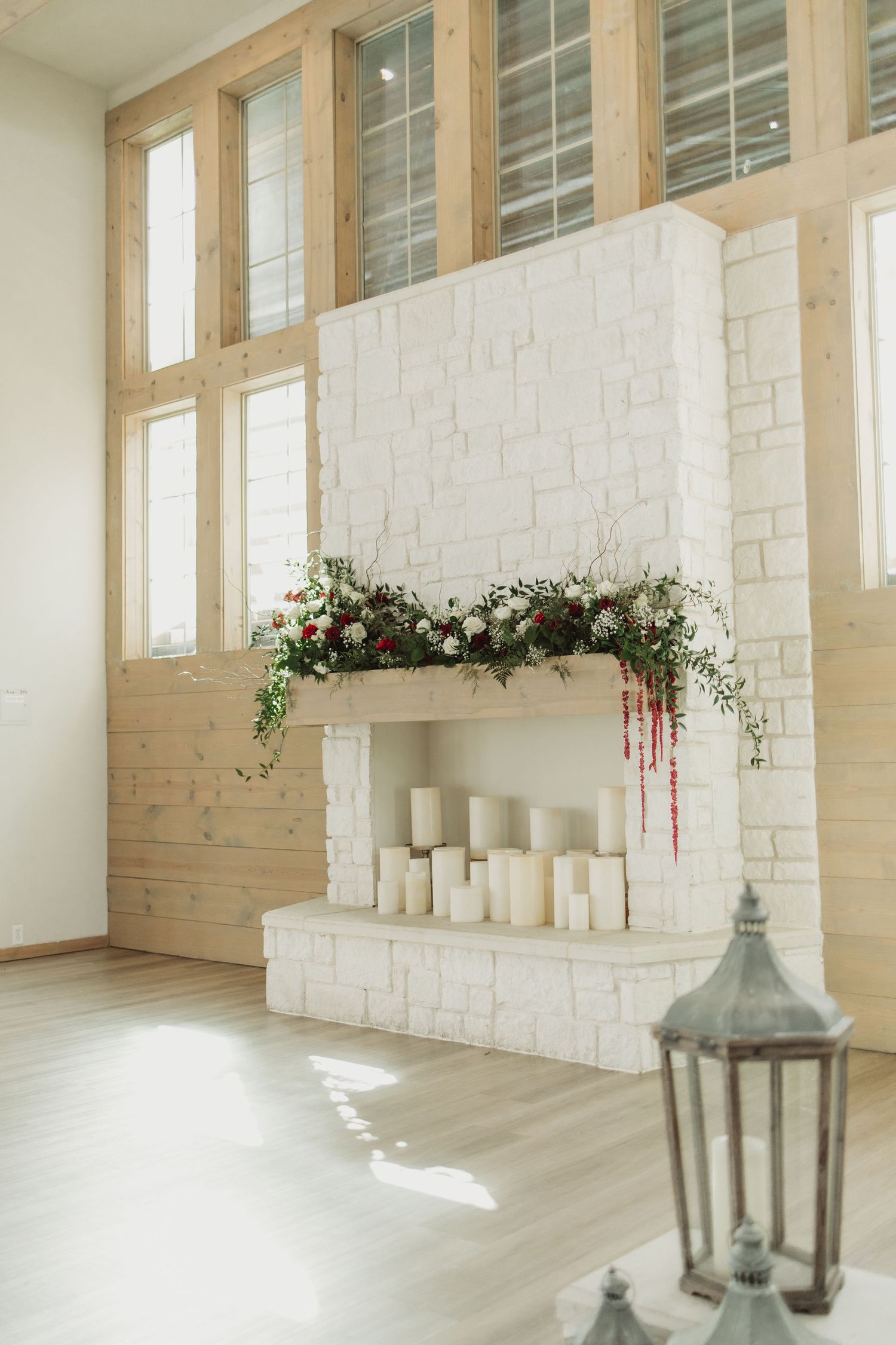 White brick fireplace decorated with floral garland and candles in a bright modern room with wood-framed windows.