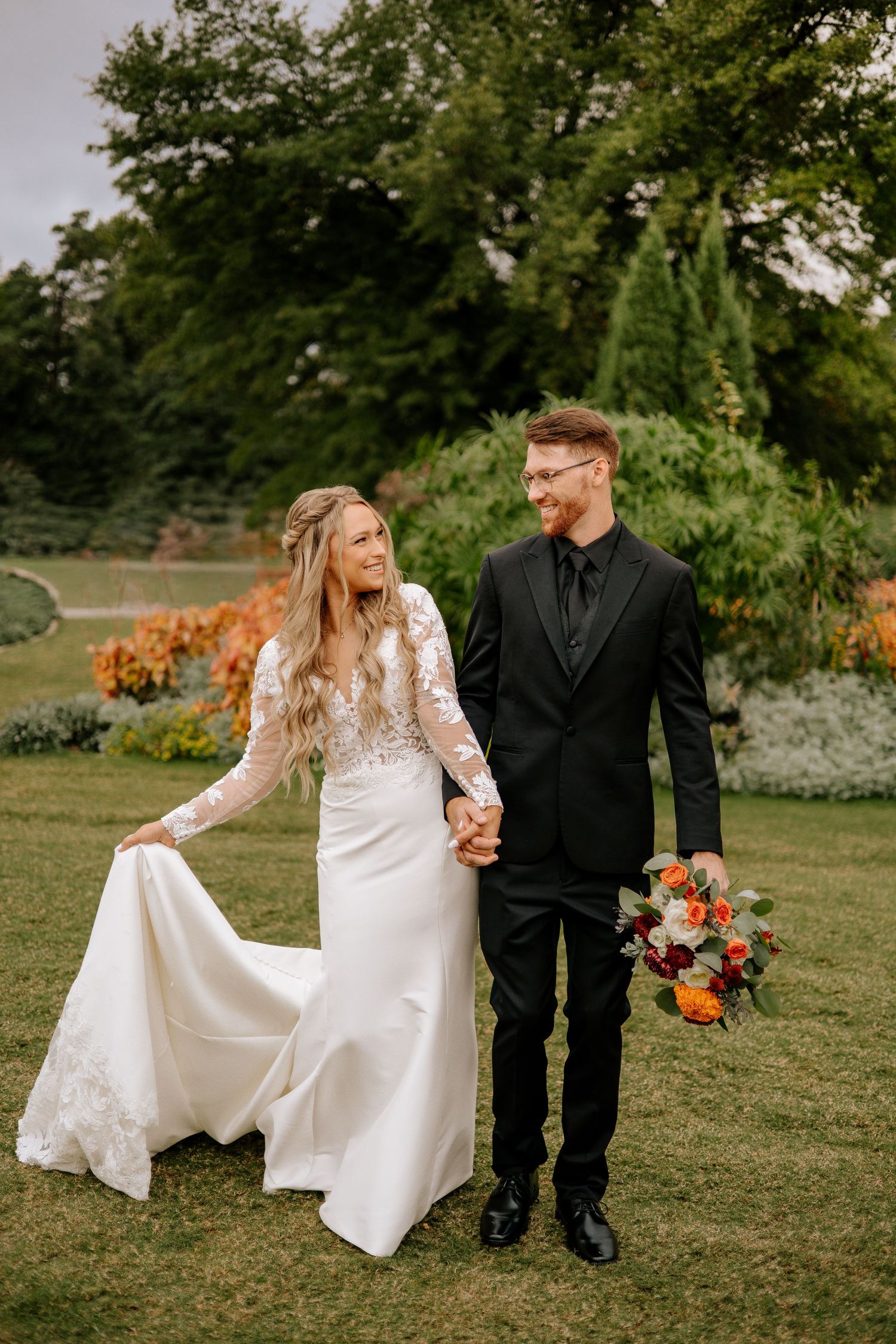 A bride and groom walk hand-in-hand through a lush garden, she in a lace gown, he in a black tuxedo holding a bouquet.