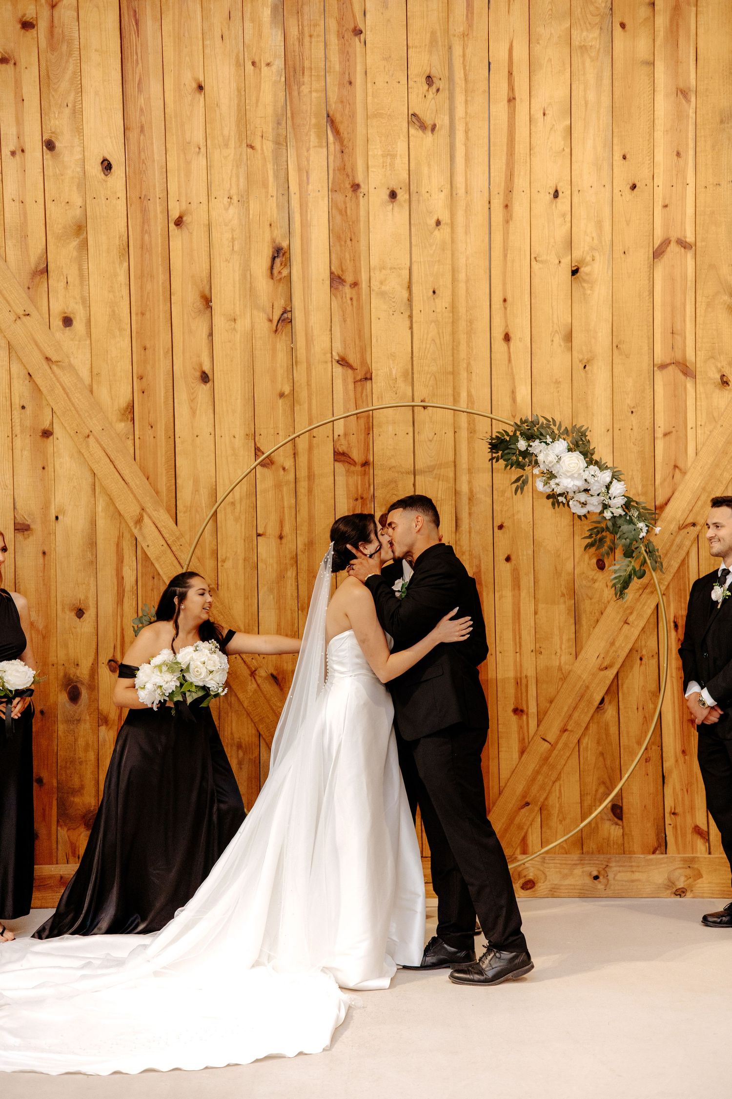 Bride and groom share first kiss under gold hoop arch at rustic barn wedding ceremony.