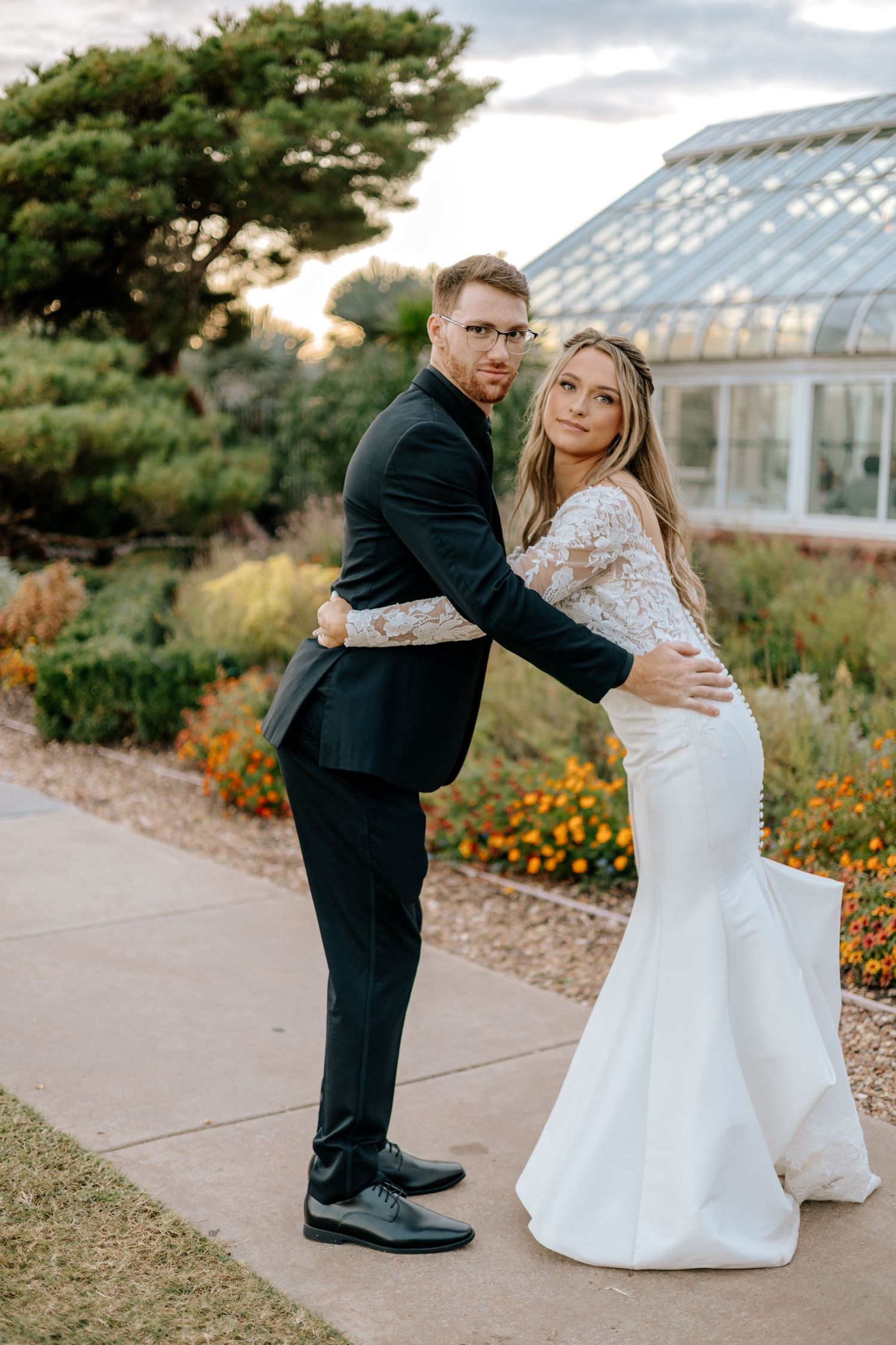 Bride and groom pose back-to-back embracing near greenhouse with colorful garden in background.