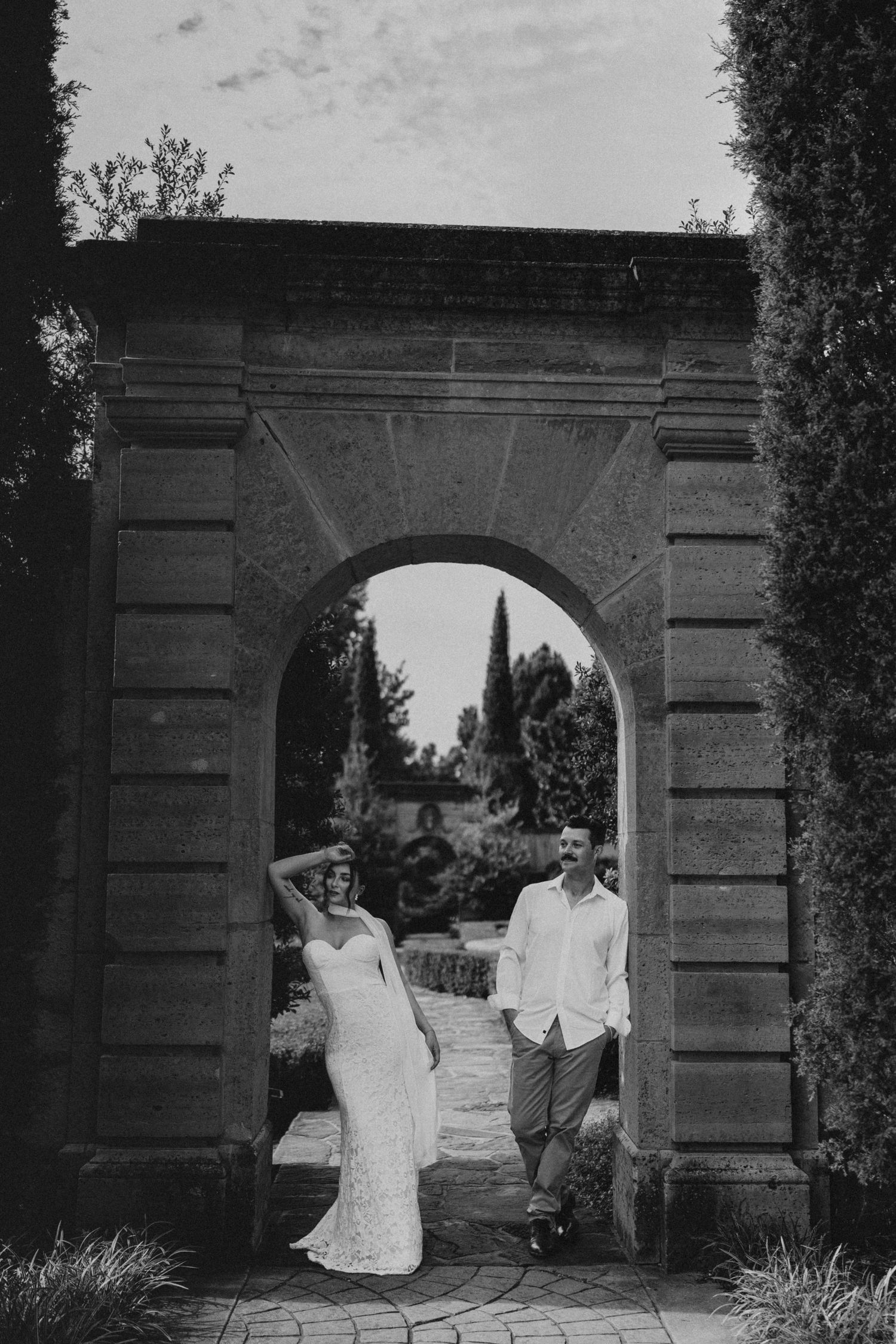Black and white photo of a bride and groom posing under a stone arch in a garden wedding setting.