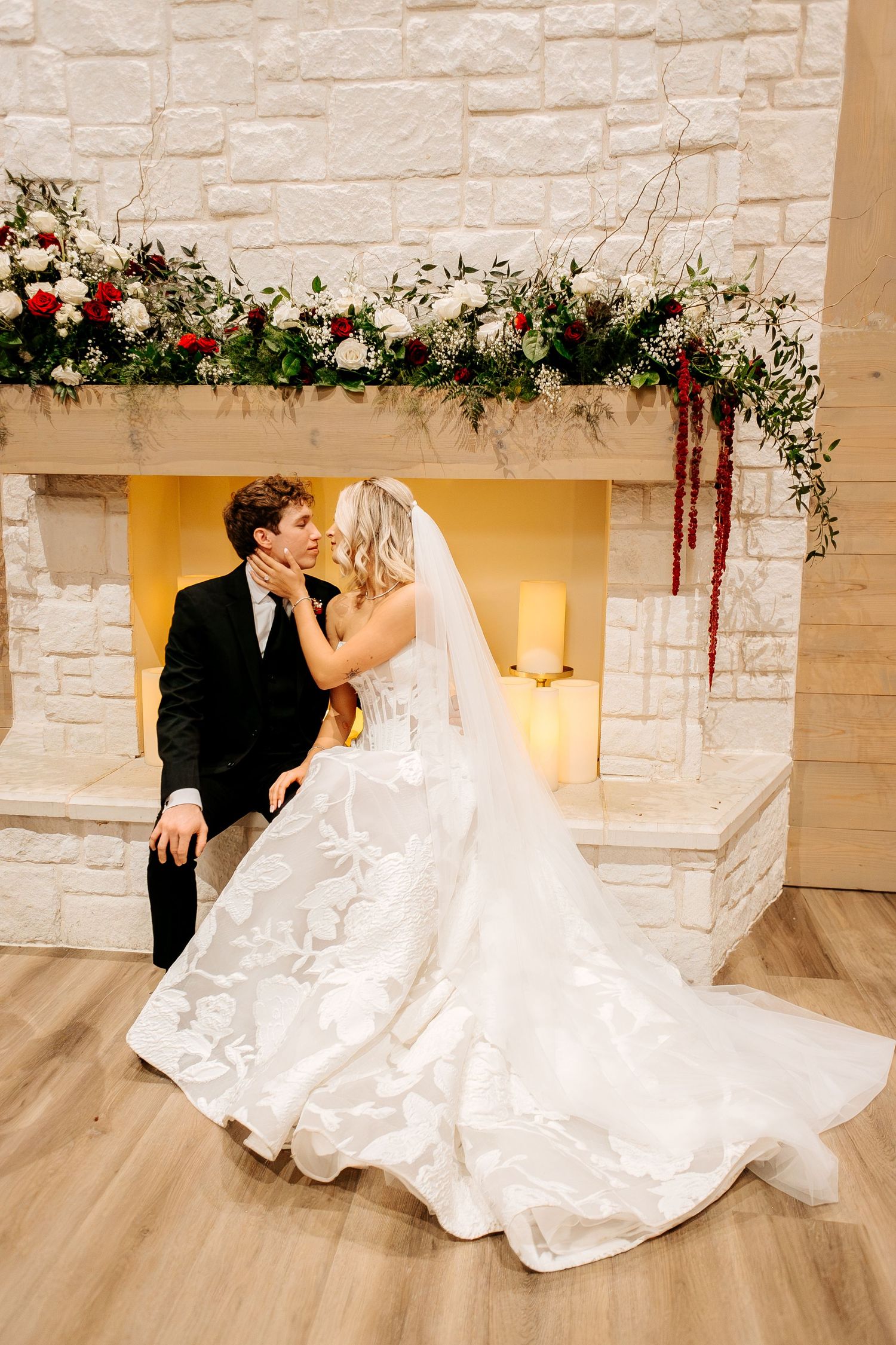 Bride and groom share romantic kiss by stone fireplace adorned with red and white floral garland.