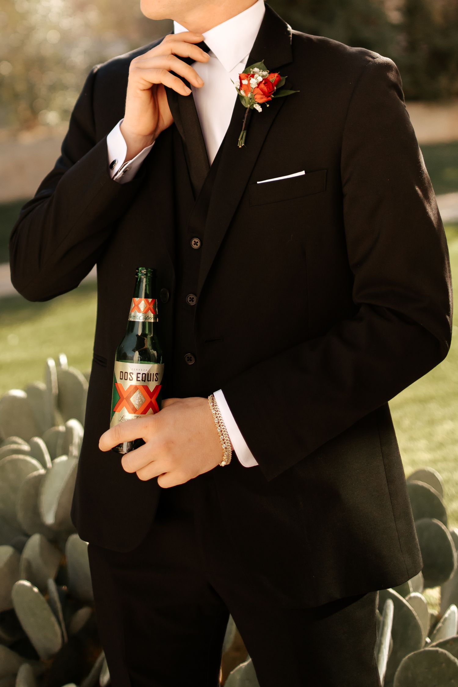 Groom in black three-piece suit adjusting tie, holding Dos Equis beer, red boutonniere, cacti background.