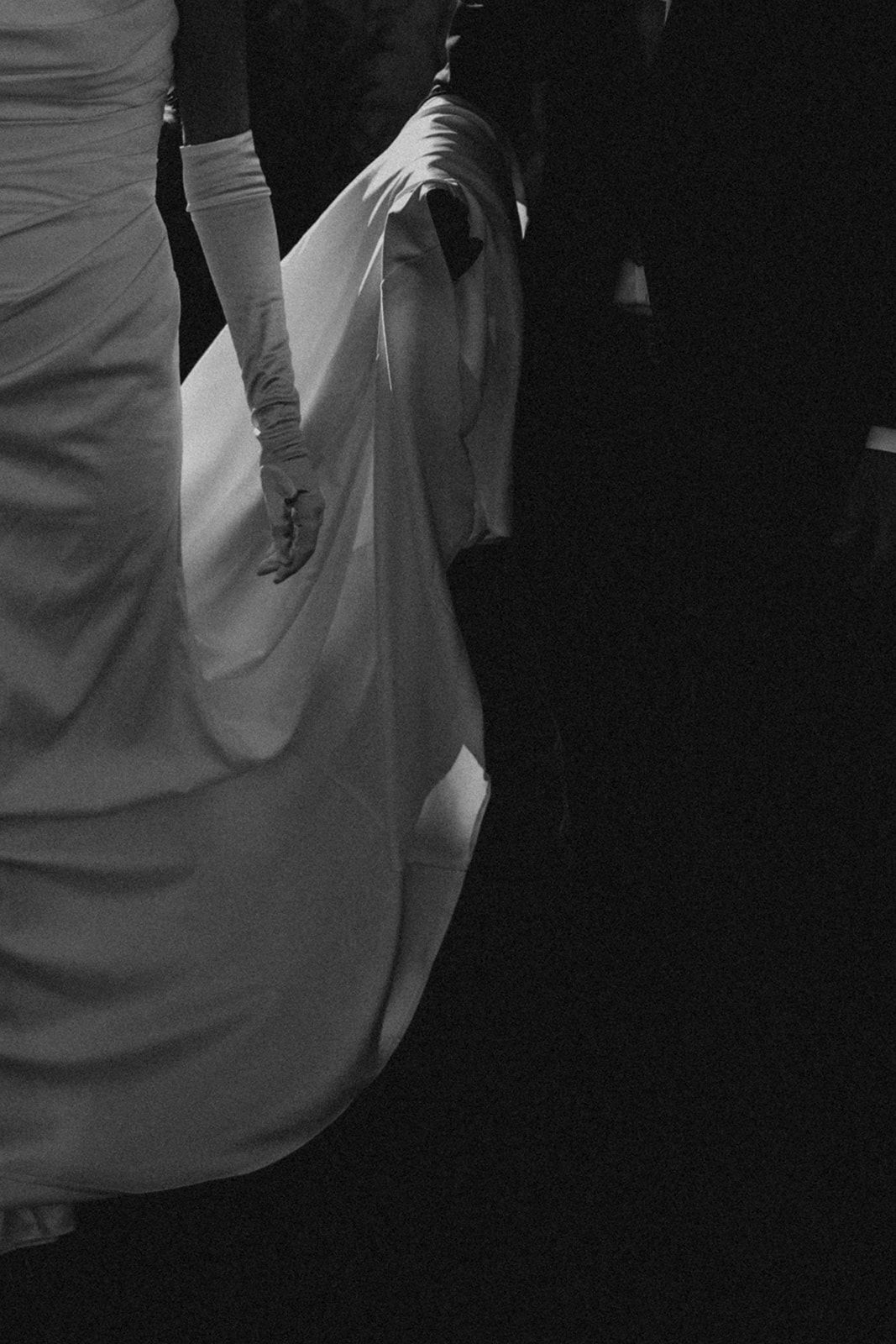 Dramatic black and white close-up of bride in white gown and gloves holding flowing train.
