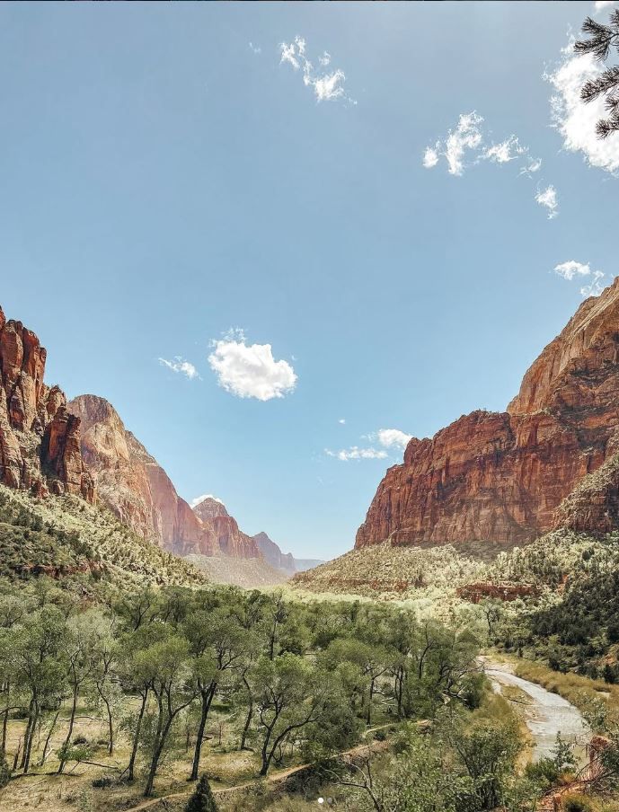 Scenic Zion National Park canyon with red sandstone cliffs, lush green trees, winding river path, and blue sky with clouds.
