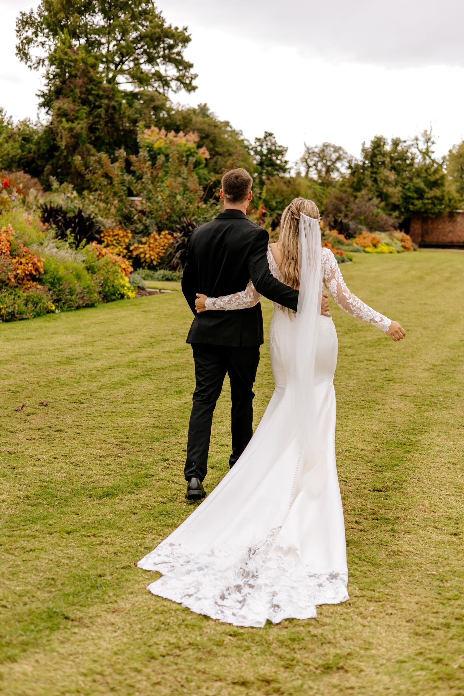 Bride and groom stroll through vibrant fall garden, bride wearing elegant fitted white gown with lace cathedral veil.