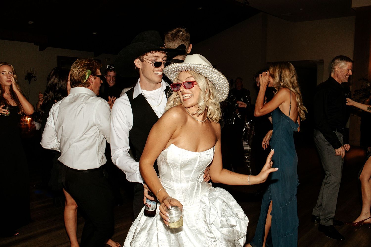 Bride in white strapless gown and hat dancing with groom in sunglasses at lively wedding reception.