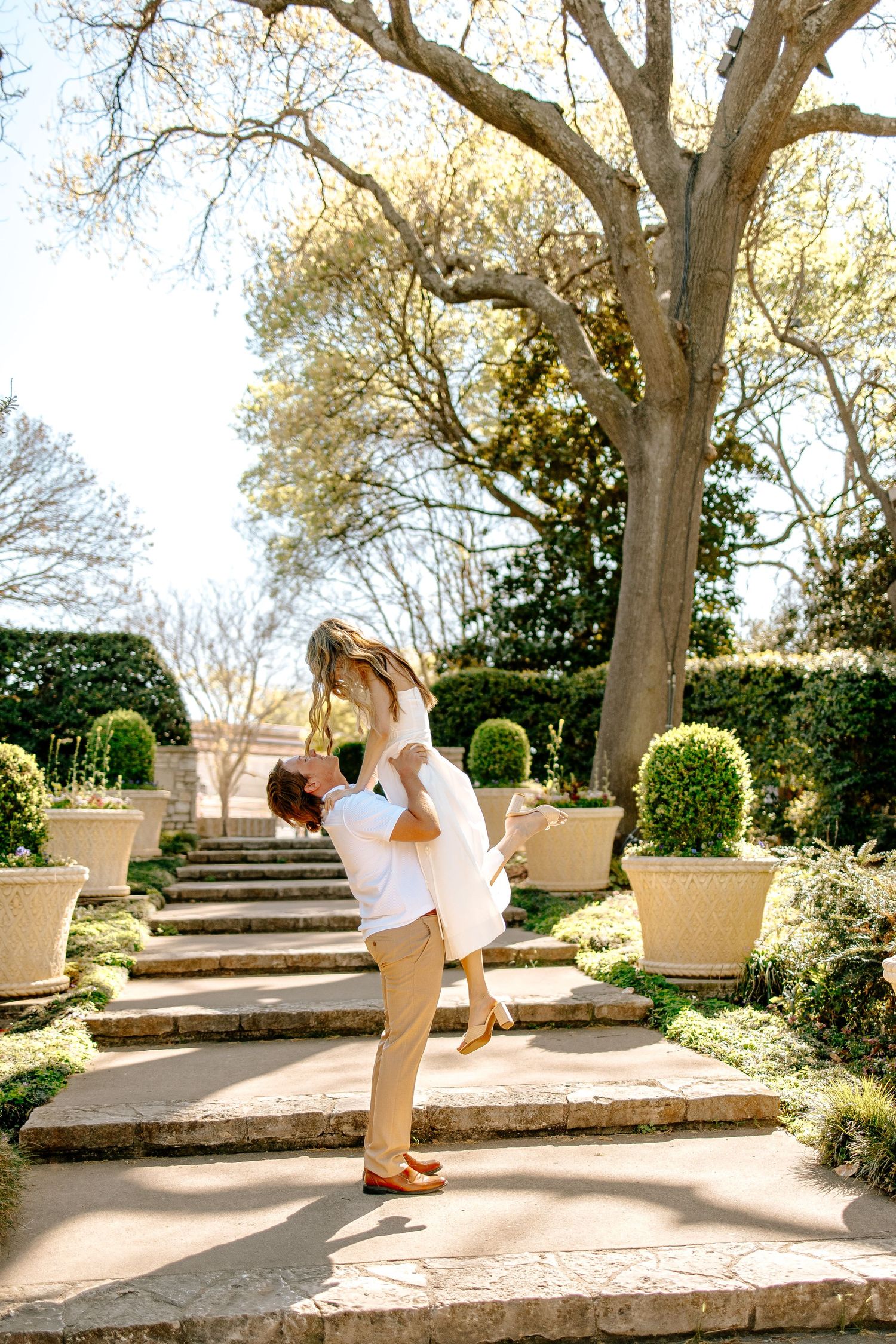 Groom lifts bride joyfully on a sunlit garden stairway surrounded by topiaries and potted plants.