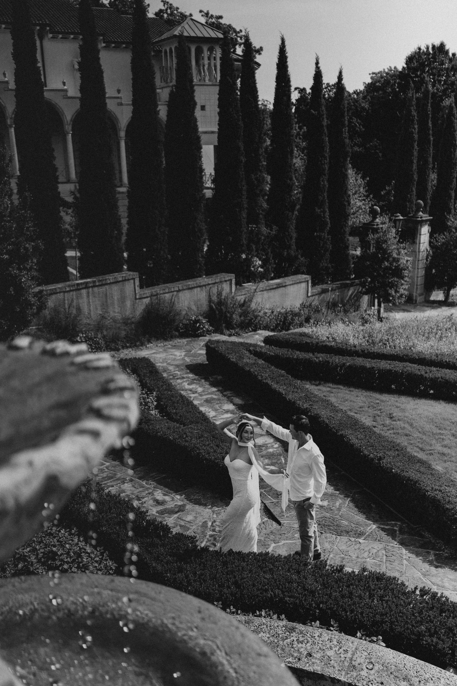 Black and white photo of a couple dancing in a formal garden with cypress trees and historic building backdrop.