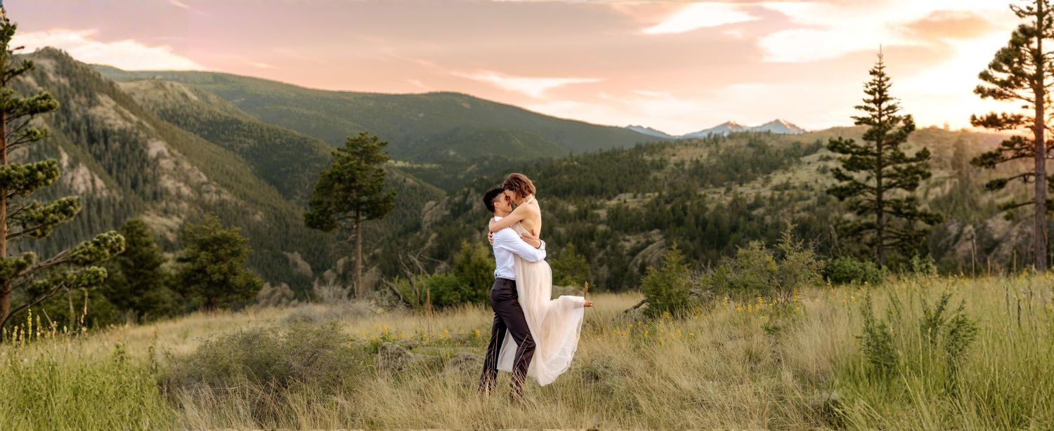 Couple embracing at sunset in a mountain meadow, with rolling hills and pine trees in the background.