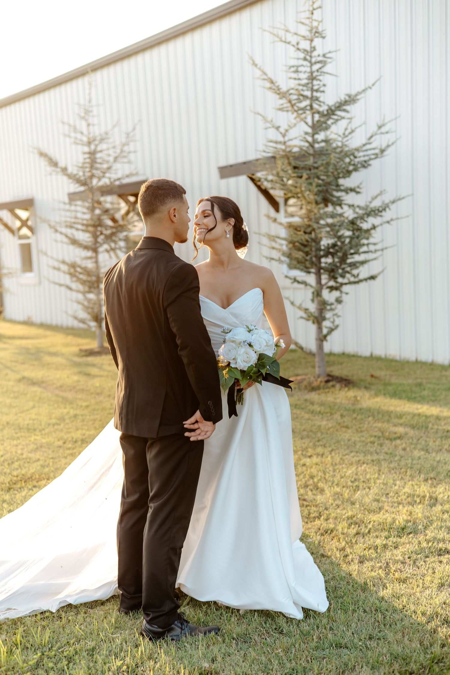 Bride in white gown and groom in black suit share romantic moment outside white barn.