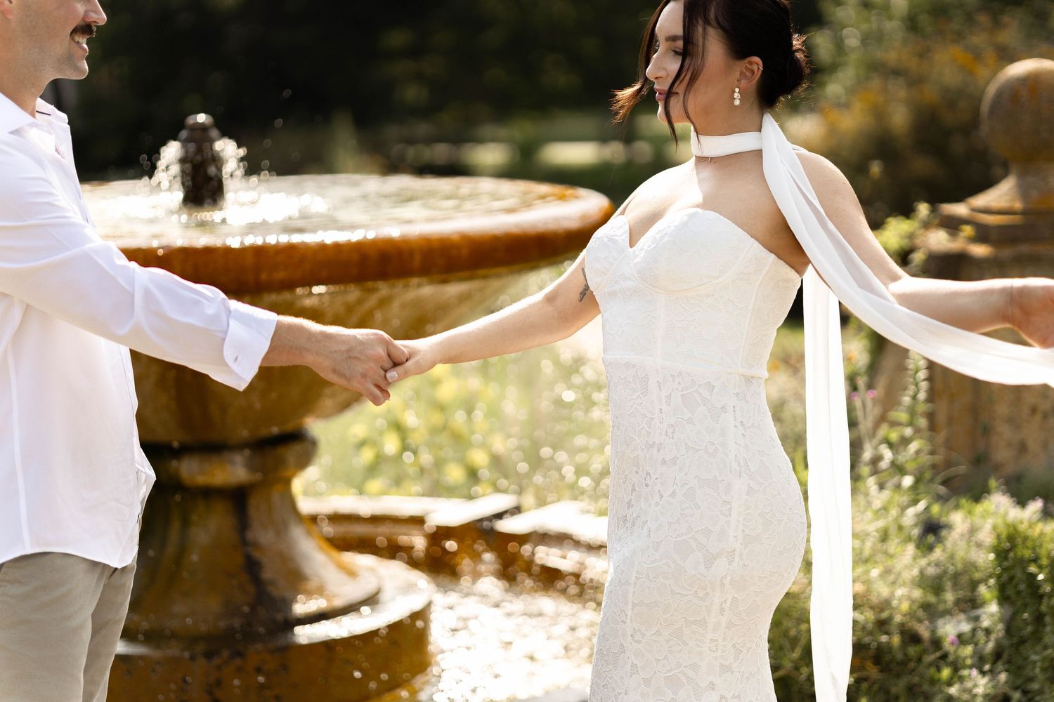 Bride in white halter gown holds groom's hand near stone fountain in sunlit garden wedding photo.