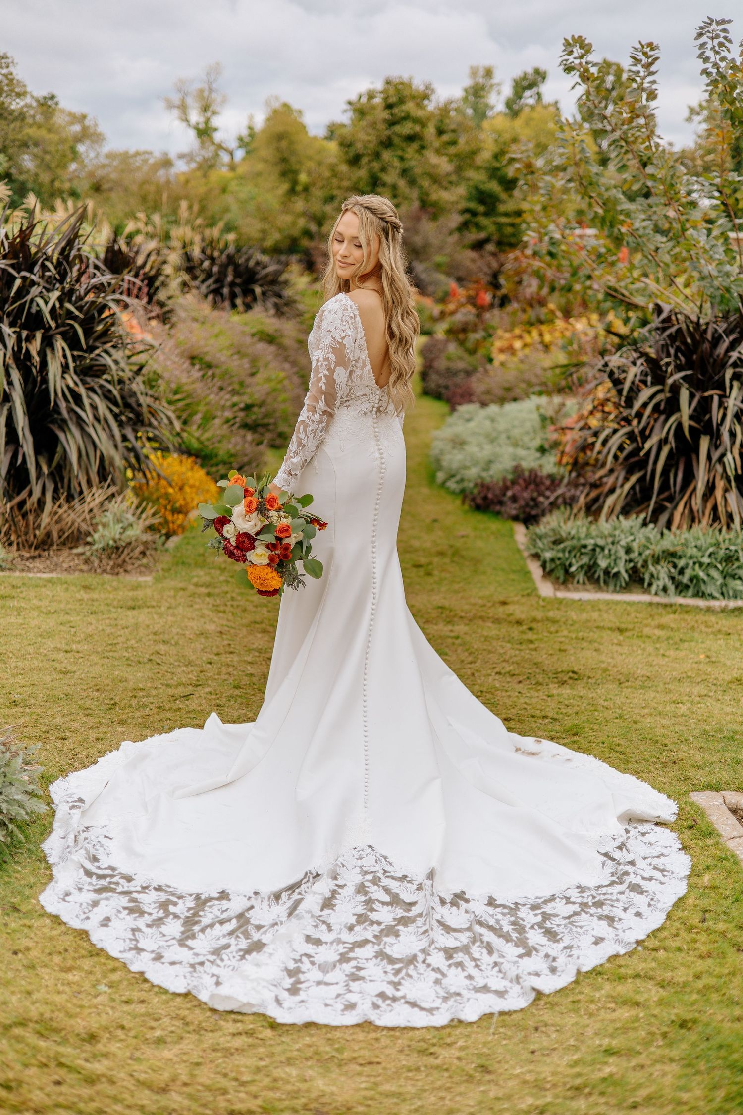 Bride in a long-sleeve lace wedding gown with an elegant lace train poses in a colorful autumn garden landscape.