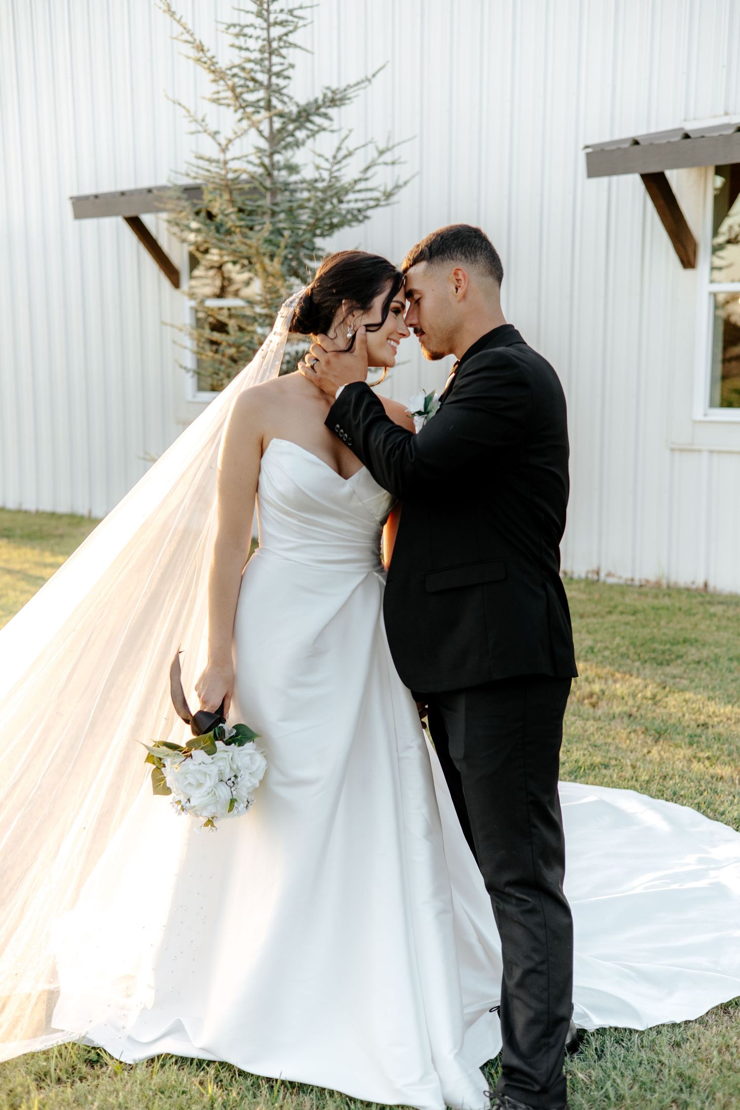 Bride and groom share an intimate forehead touch moment outside a white barn venue at golden hour.