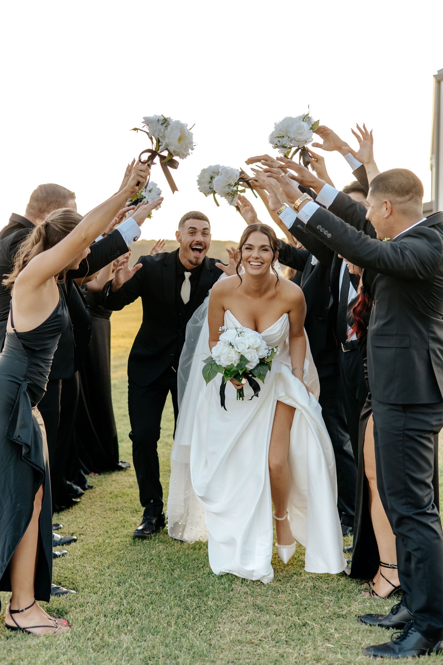 Bride and groom walk through cheering wedding party tunnel holding white bouquets outdoors.