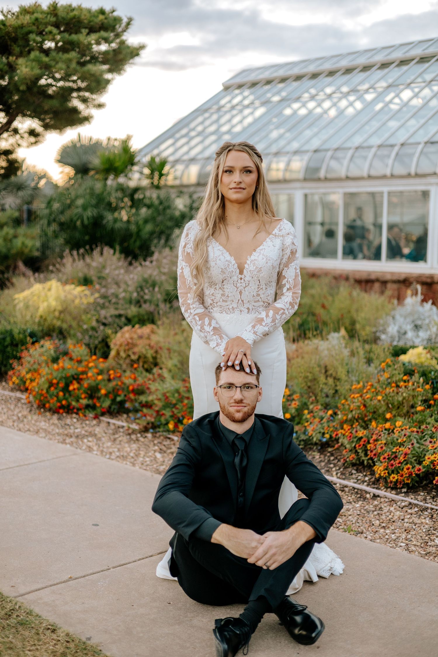 Bride stands behind seated groom in elegant portrait outside glass greenhouse with orange flowers.