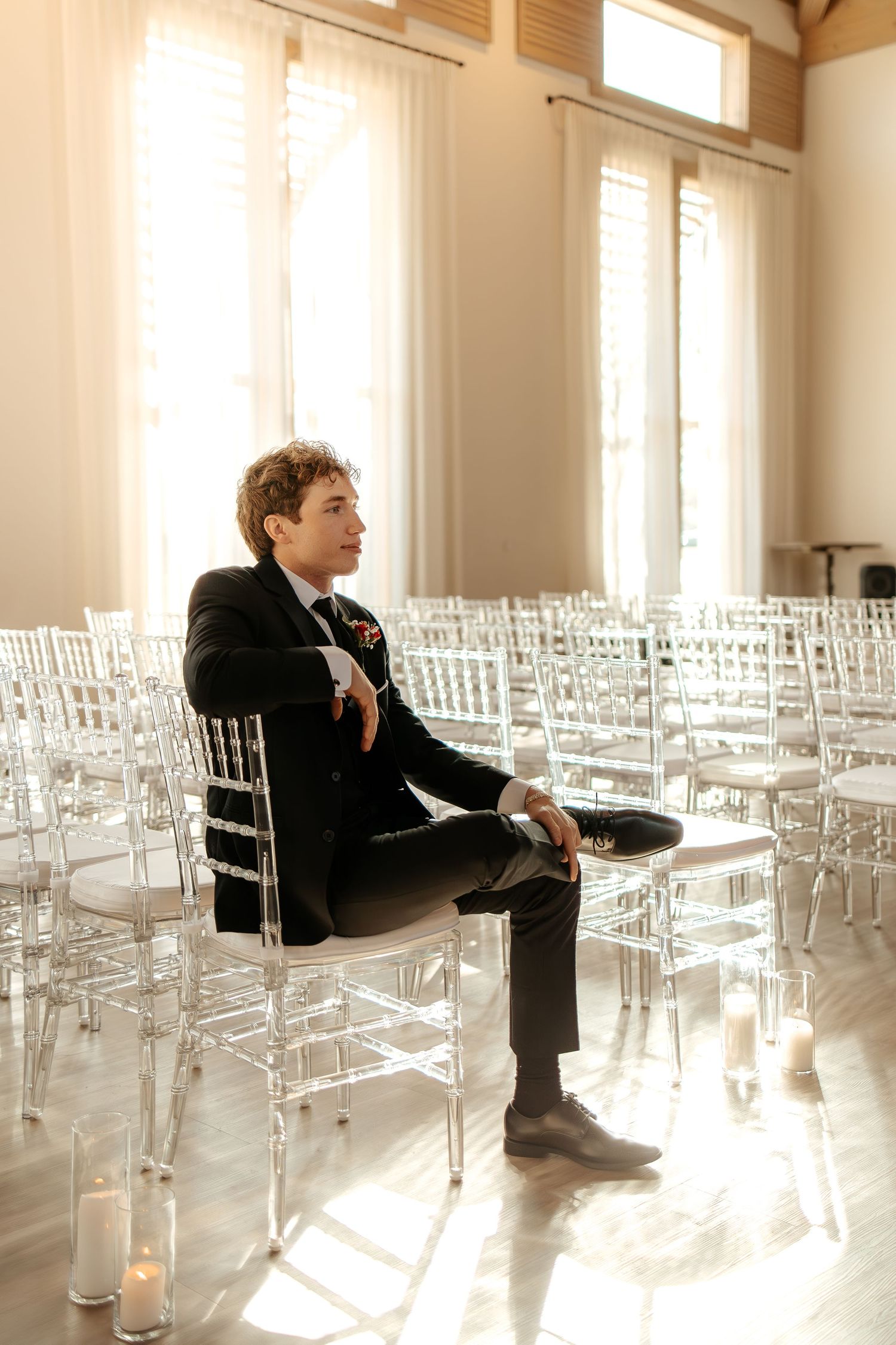 Dapper groom in black tuxedo sits alone among clear chiavari chairs in sunlit wedding ceremony hall.