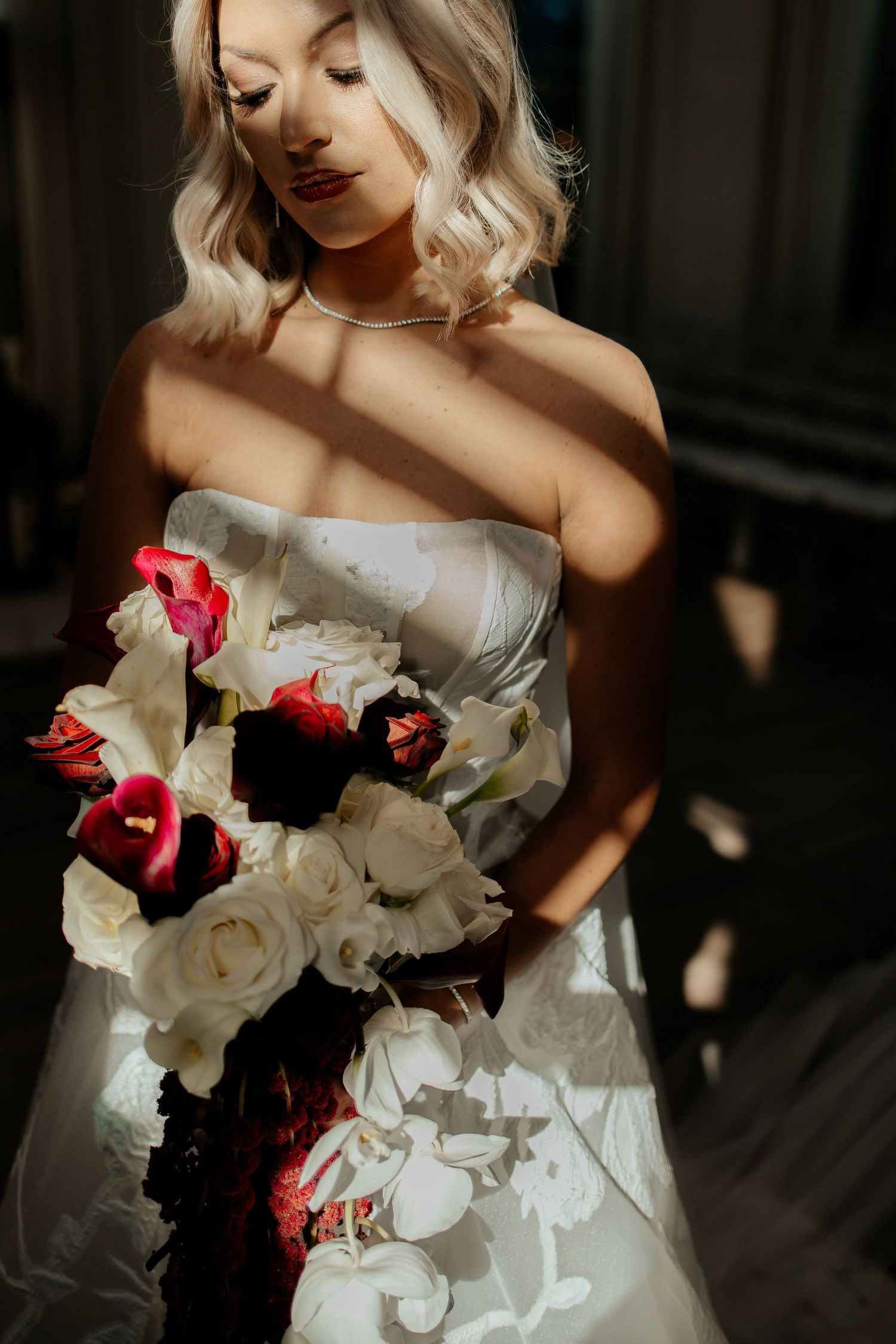 Blonde bride in strapless gown holds dramatic red and white bouquet in moody natural lighting.