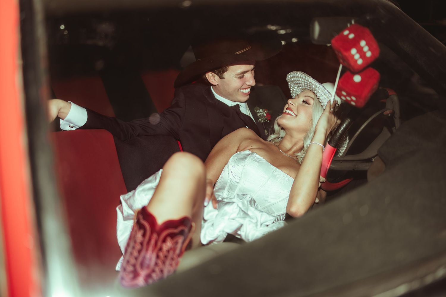 Bride in white dress and red cowboy boots laughs with groom inside a vintage red car.