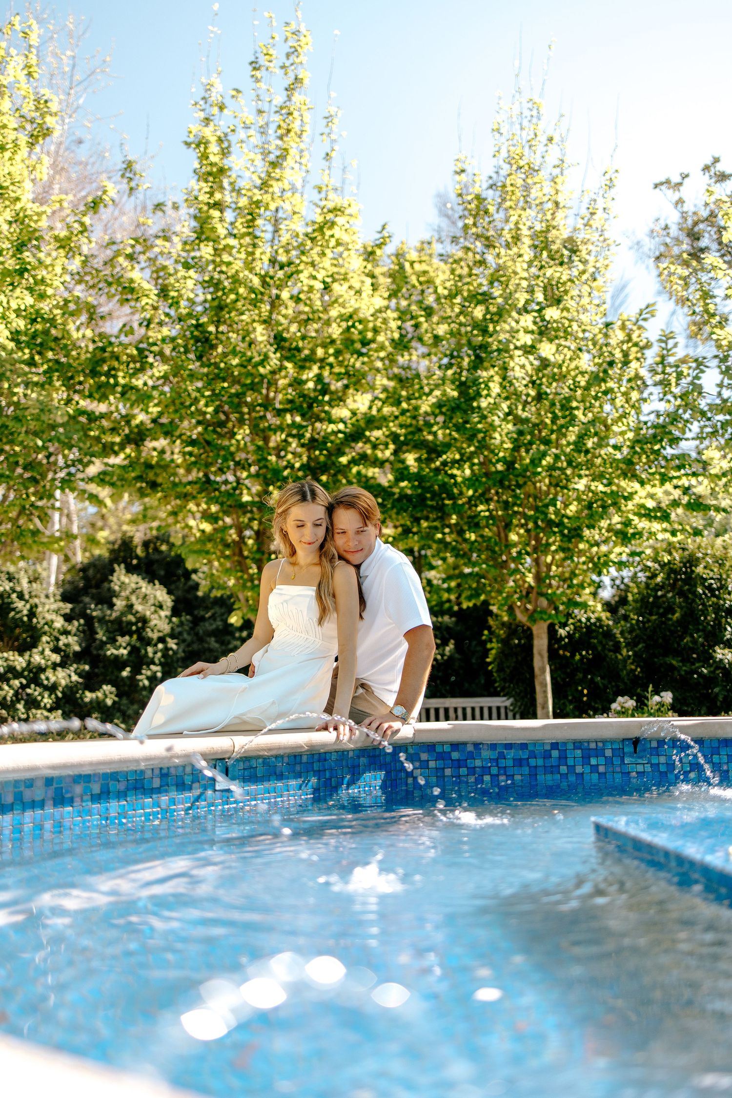 Engaged couple sitting together by a blue mosaic pool surrounded by lush green trees outdoors.